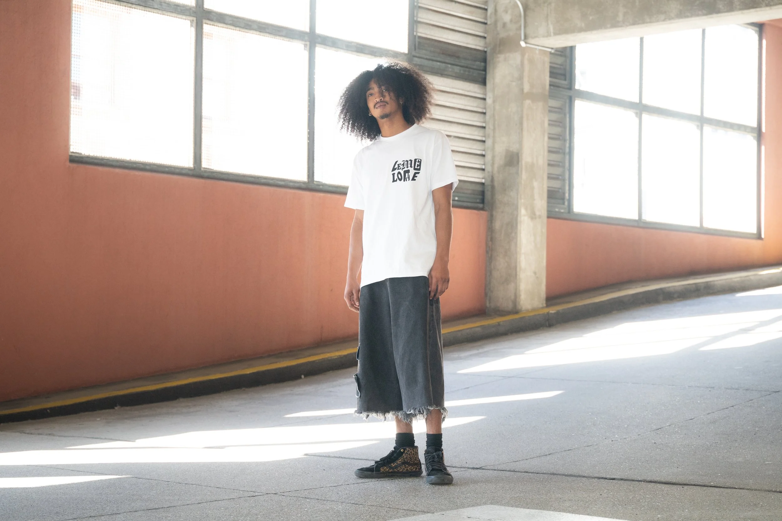 A young man with curly hair and a mustache standing in a parking garage with large windows, wearing a white T-shirt with black text, black socks, and leopard print high-top sneakers.