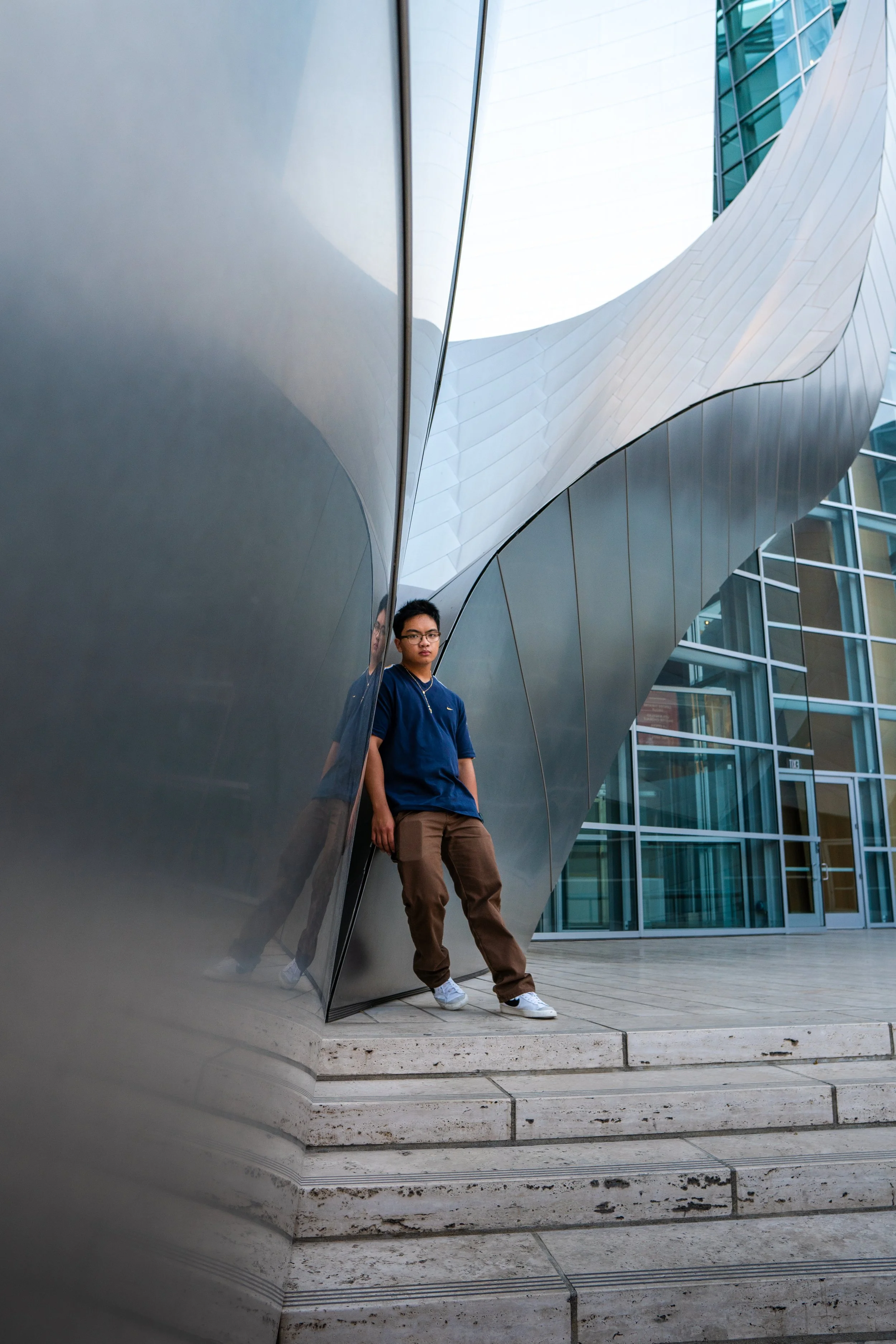 A young man wearing glasses, a blue shirt, brown pants, and white sneakers leaning against a reflective, modern metal structure outside a glass building.