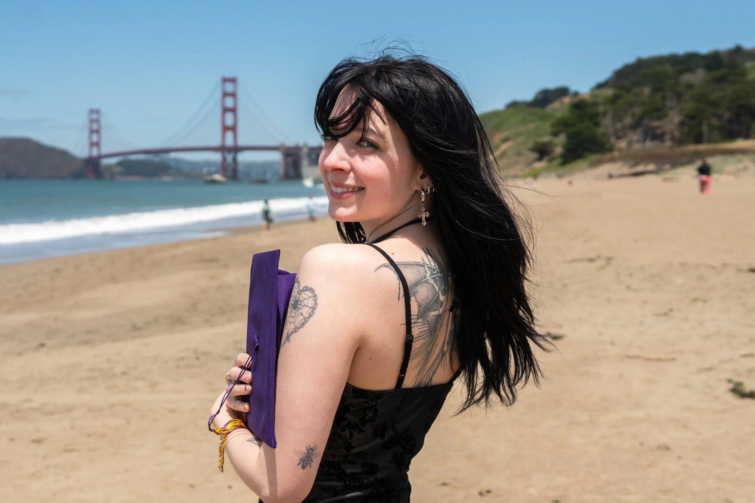 A woman with black hair, earrings, and tattoos on her arm and shoulder smiling on a sandy beach, with the Golden Gate Bridge, blue sky, and ocean in the background.