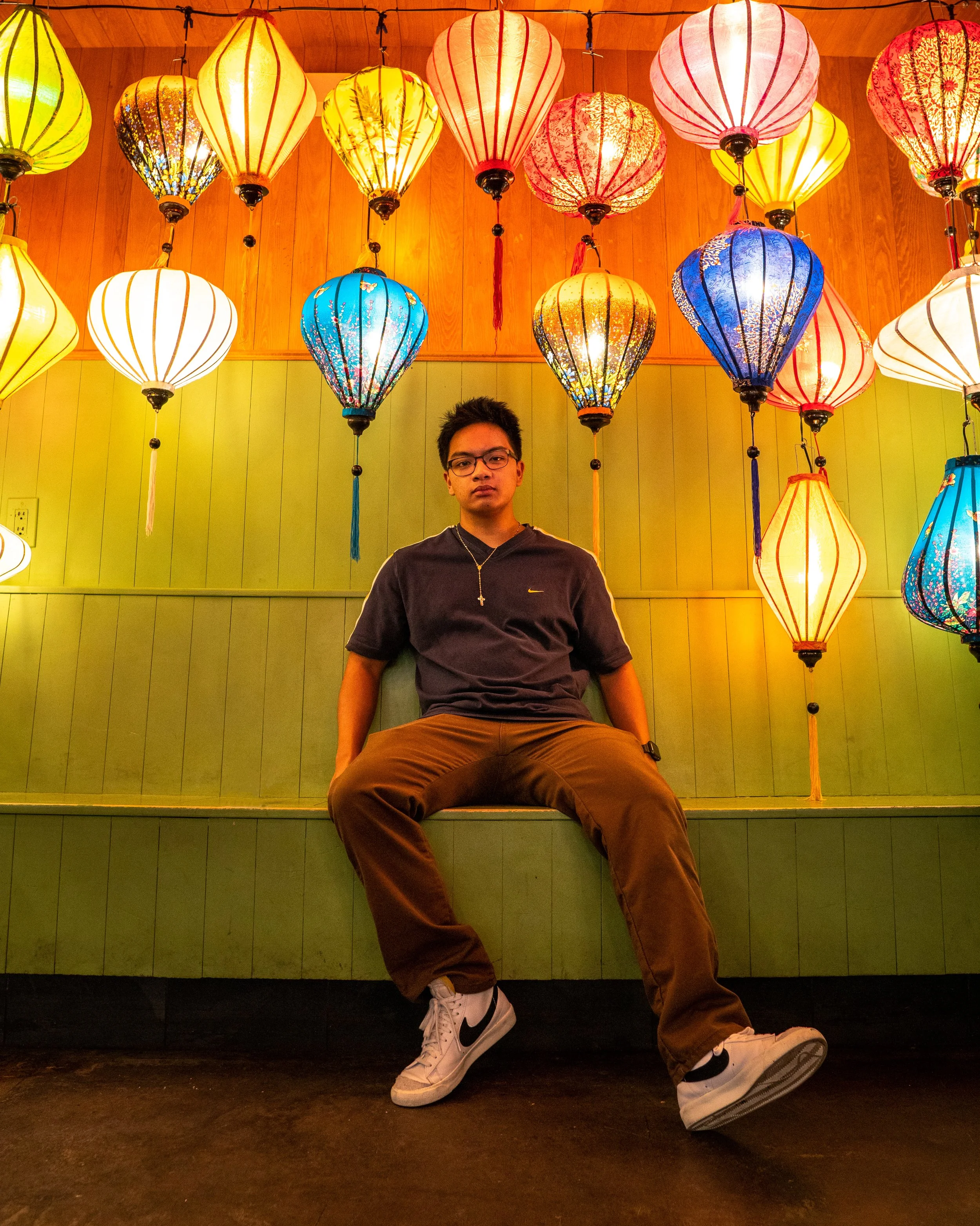 A young man sitting on a green bench surrounded by colorful hanging paper lanterns at night.