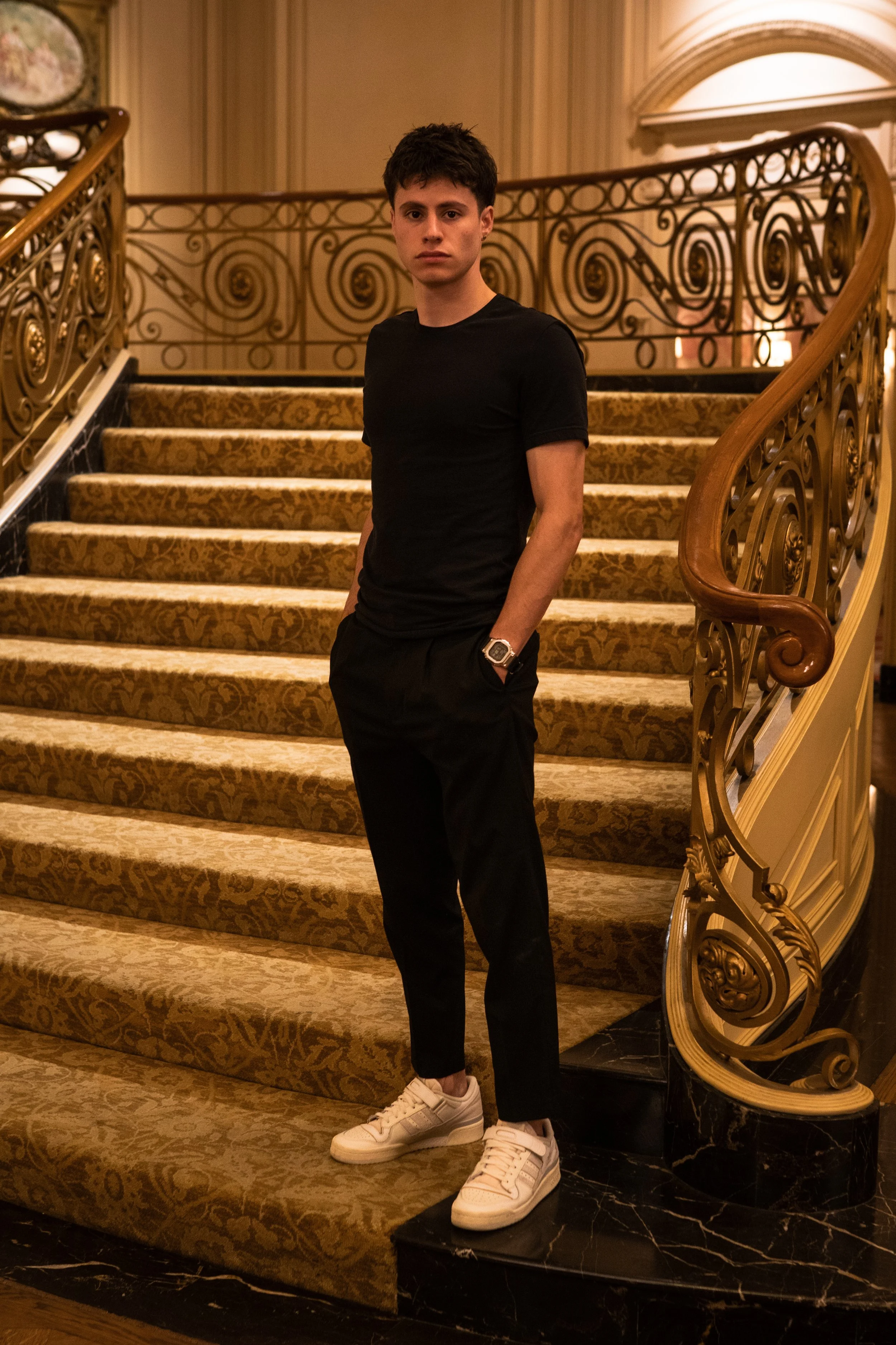 Young man standing on a staircase inside an ornate, luxurious hotel or mansion, wearing a black t-shirt, black pants, white sneakers, and a watch, with a serious expression.