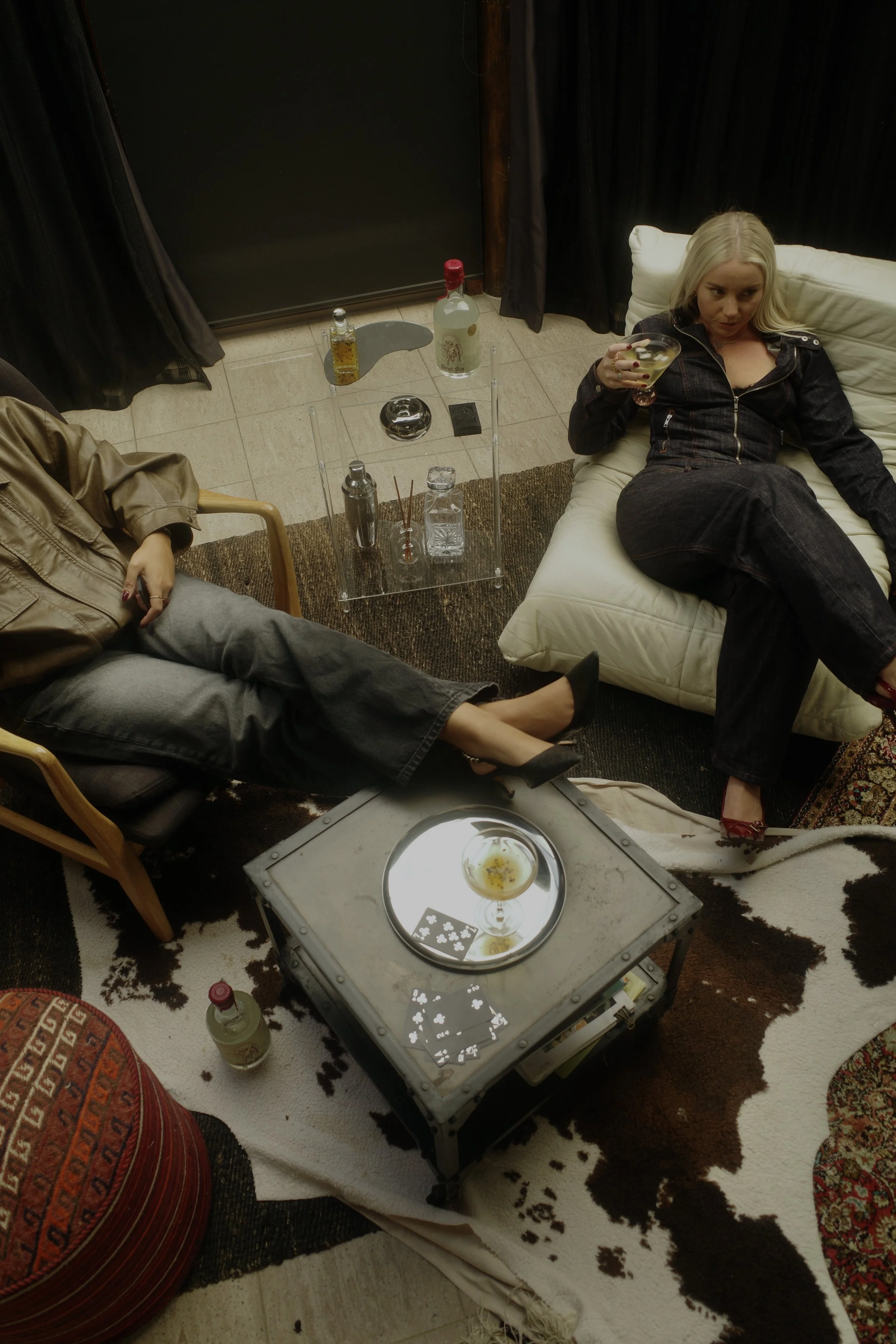 Two women relax in a living room, one on a chair and the other on a sofa, with drinks and playing cards on a reflective tray table.
