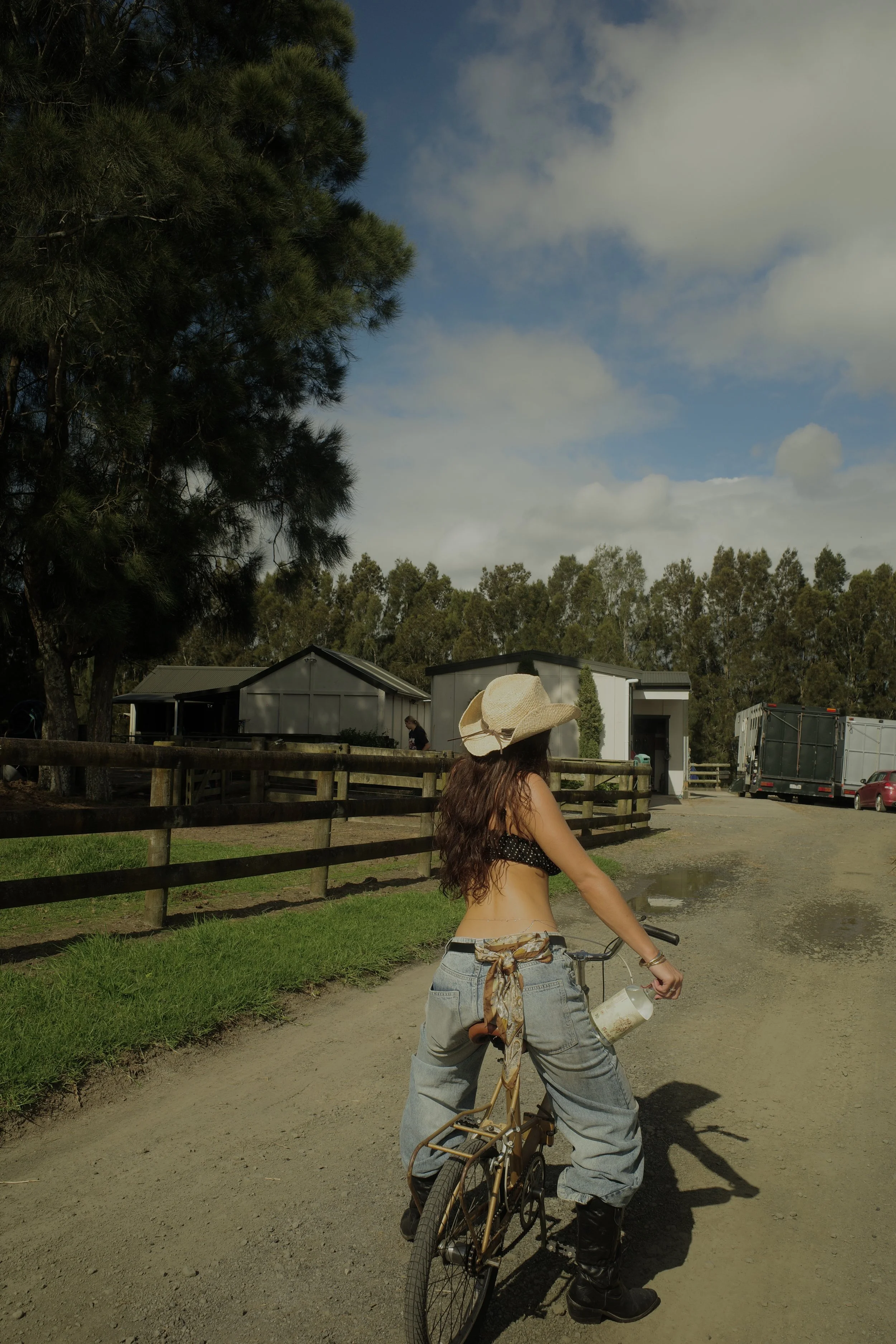 A woman with long brown hair, wearing a large straw hat, a black polka dot crop top, baggy jeans with a tie around the waist, and black boots, stands beside a vintage bicycle on a dirt path. She is outdoors in a rural or farm setting with green grass, tall trees, a wooden fence, and several buildings, with a partly cloudy sky above.