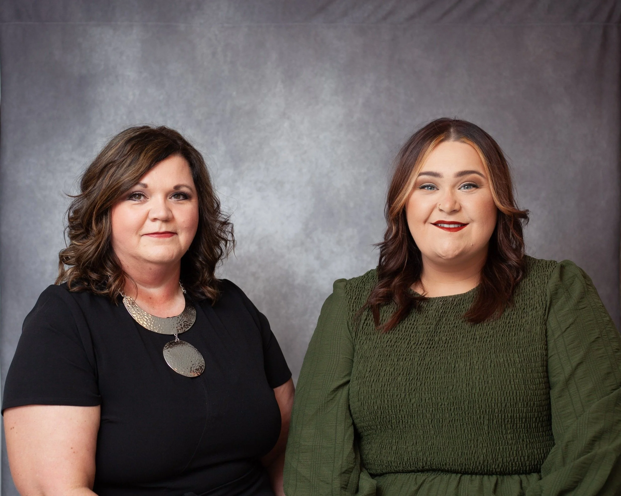 Two women sitting together, one wearing a black shirt and a large metallic necklace, the other in a green textured top, both smiling at the camera against a gray background.