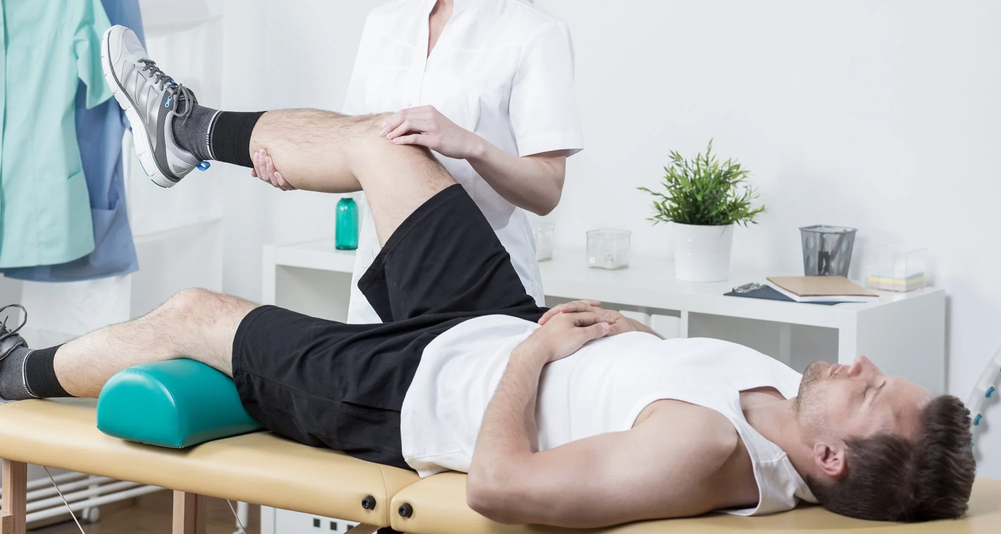 A physical therapist or doctor assisting a patient with leg exercises while lying on a treatment table in a medical office. The patient is holding his arm across his chest, and the therapist is holding his leg and checking his knee. The room has medical supplies, a plant, and paperwork on a side table.