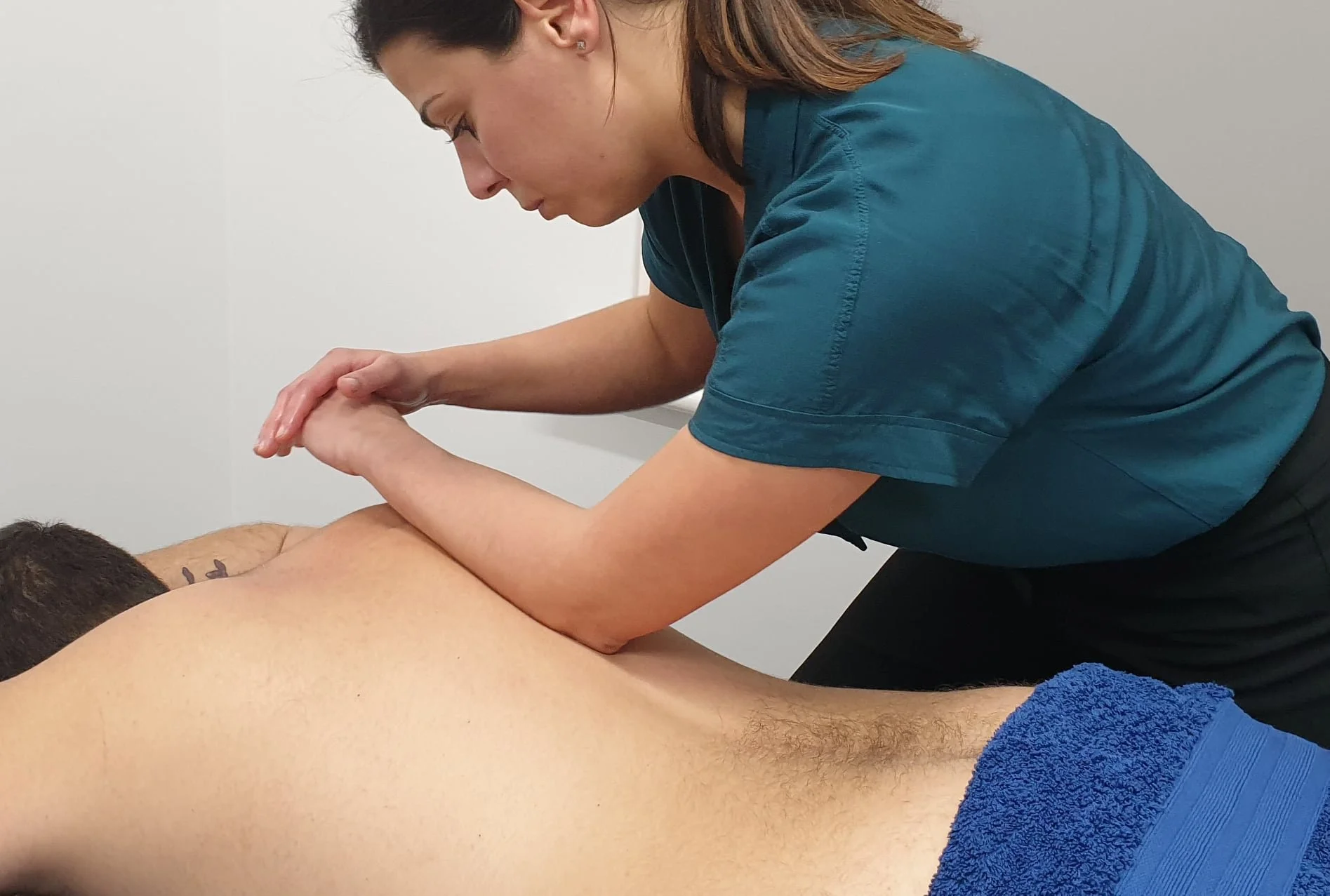A woman wearing teal scrubs gives a massage to a man lying face down on a massage table, with a blue towel covering part of his lower body, against a plain white wall background.