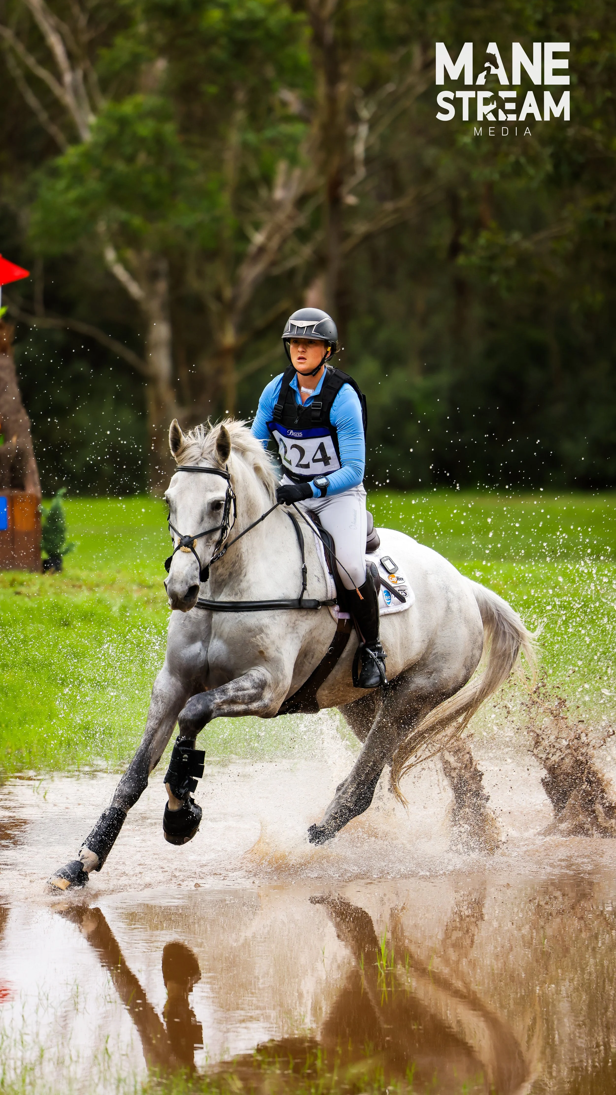 A person riding a white horse through a water obstacle during an equestrian event, wearing a helmet, blue jacket, white riding pants, and a numbered vest.