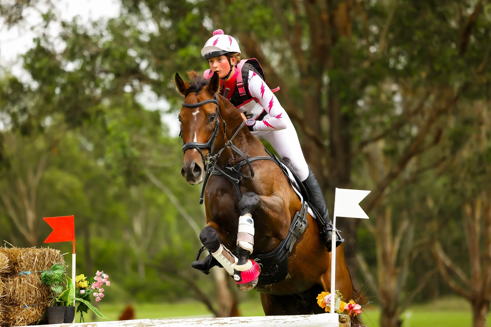 A female equestrian wearing a white helmet and outfit with pink lightning bolt patterns, riding a brown horse during a cross-country event. They are jumping over a hay bale decorated with flowers, with small flags on either side, through a wooded area.