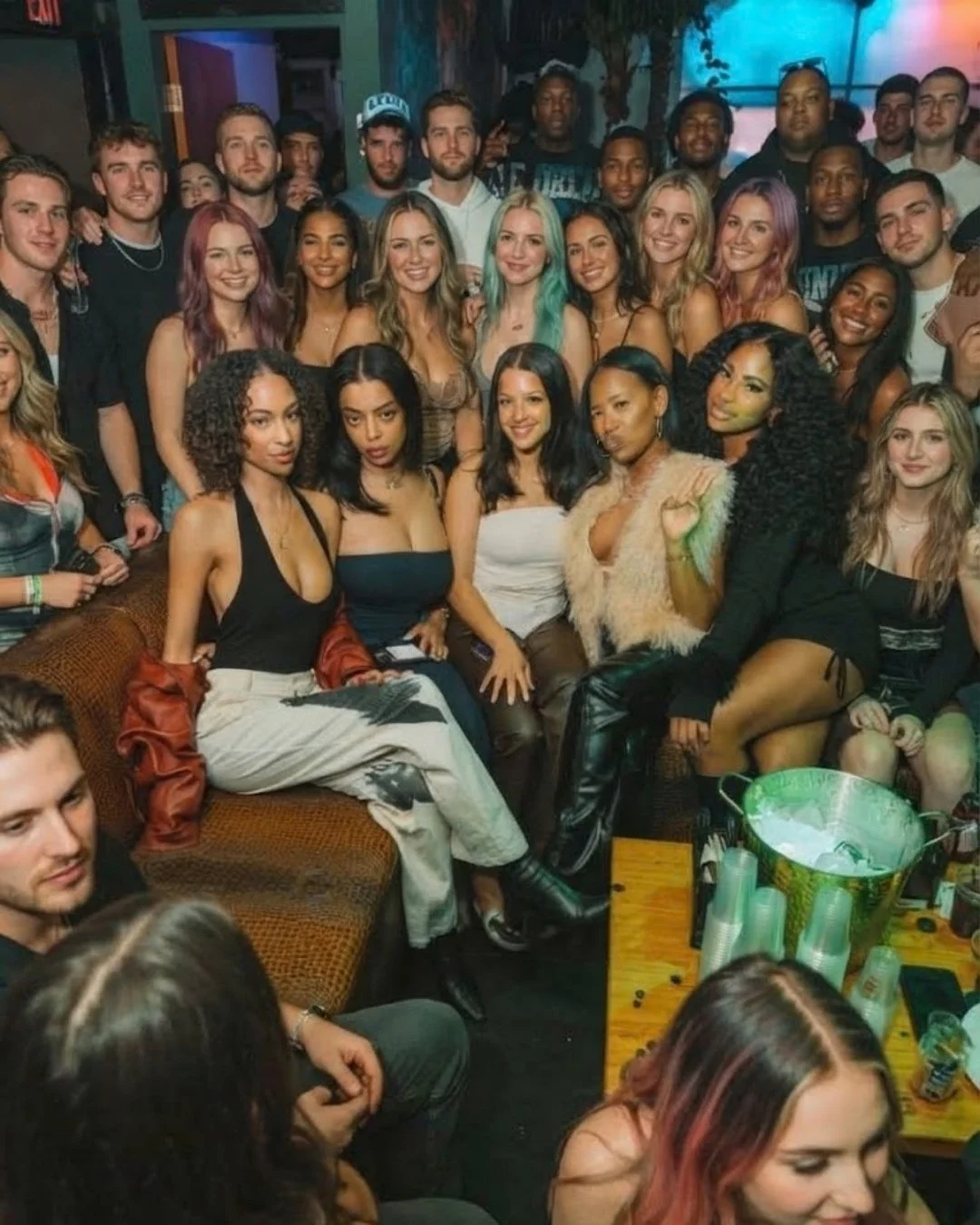 Group of young adults in a lively social setting, some seated on a couch, others standing behind, smiling and posing for the photo, with colorful lighting and a party atmosphere.