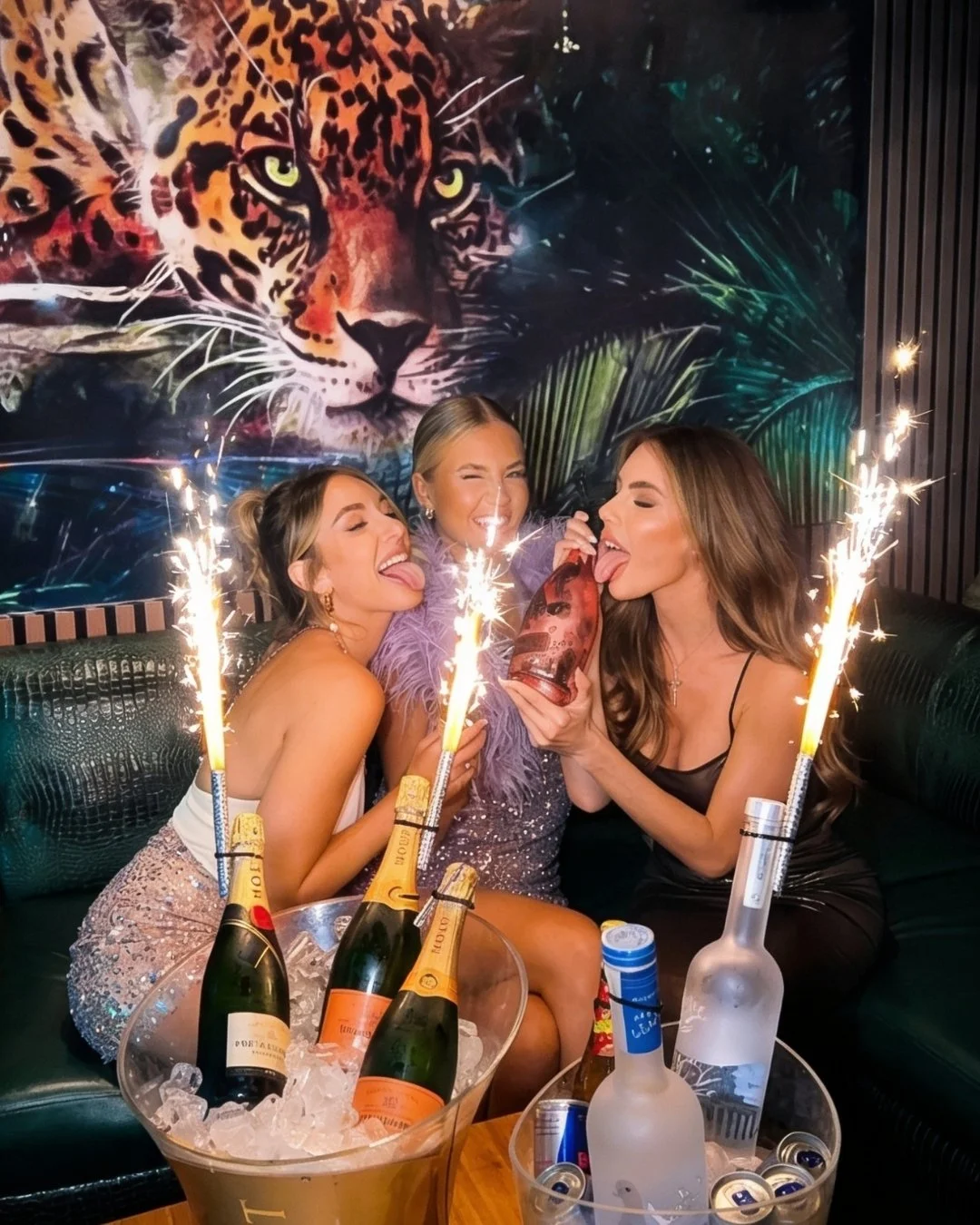Three women celebrating with champagne, sparklers, and a birthday cake with sparklers, in front of a large leopard mural.