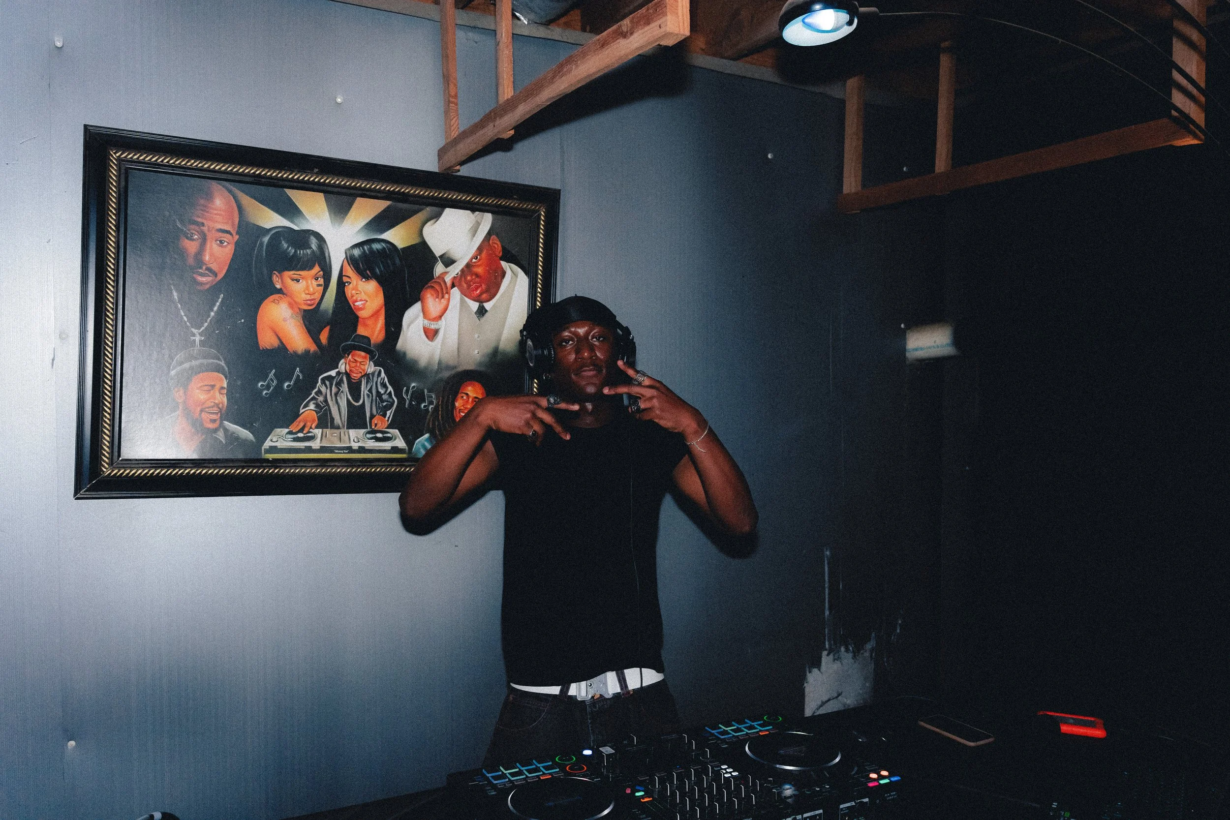 DJ wearing headphones making peace signs with both hands, standing behind a DJ mixer and turntables in a dark room with a framed painting of musicians and singers on the wall behind him.