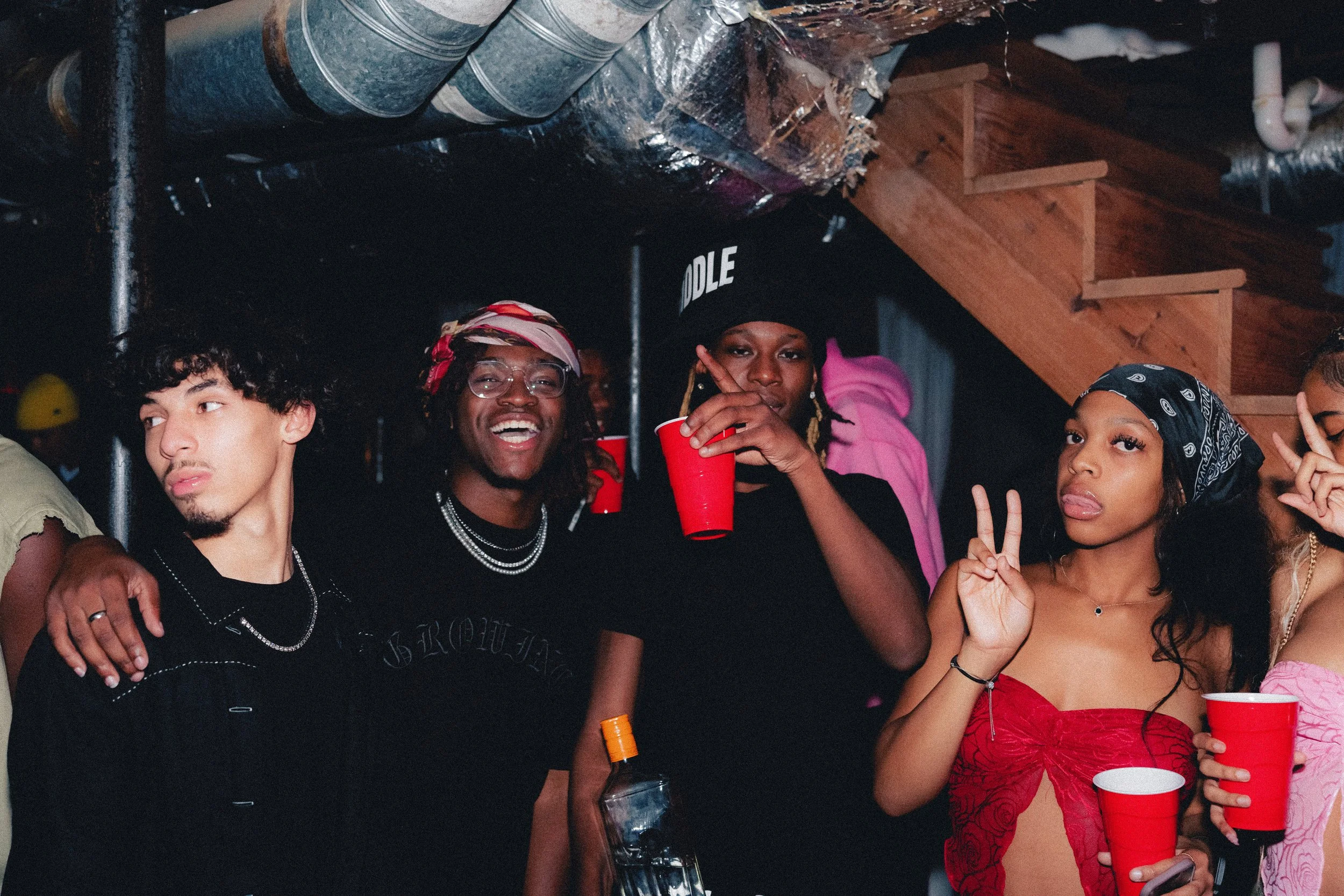 Group of young adults at a party, holding red cups, with a wooden staircase in the background, dressed in casual and trendy clothing, some making peace signs.