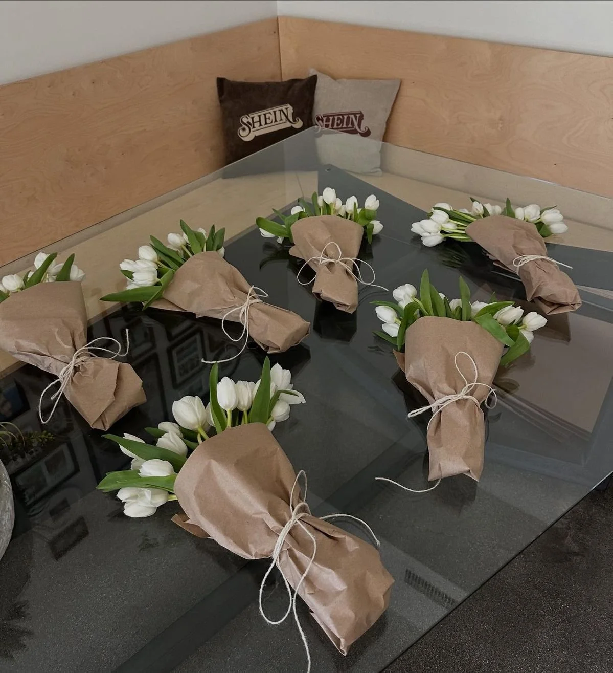 Six bouquets of white tulips wrapped in brown paper tied with string, arranged on a black glass table. In the background, there are two pillows with "SHEIN" logos on a wooden headboard.