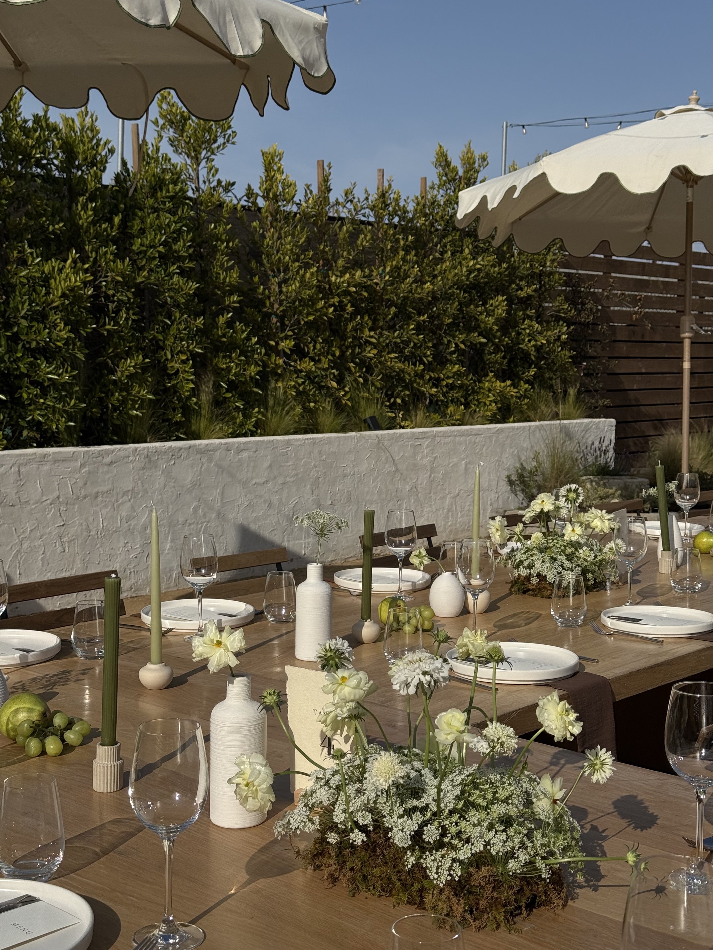 Outdoor dining table set with white plates, wine glasses, green candles, white vases, and floral centerpieces under umbrellas in a garden setting.