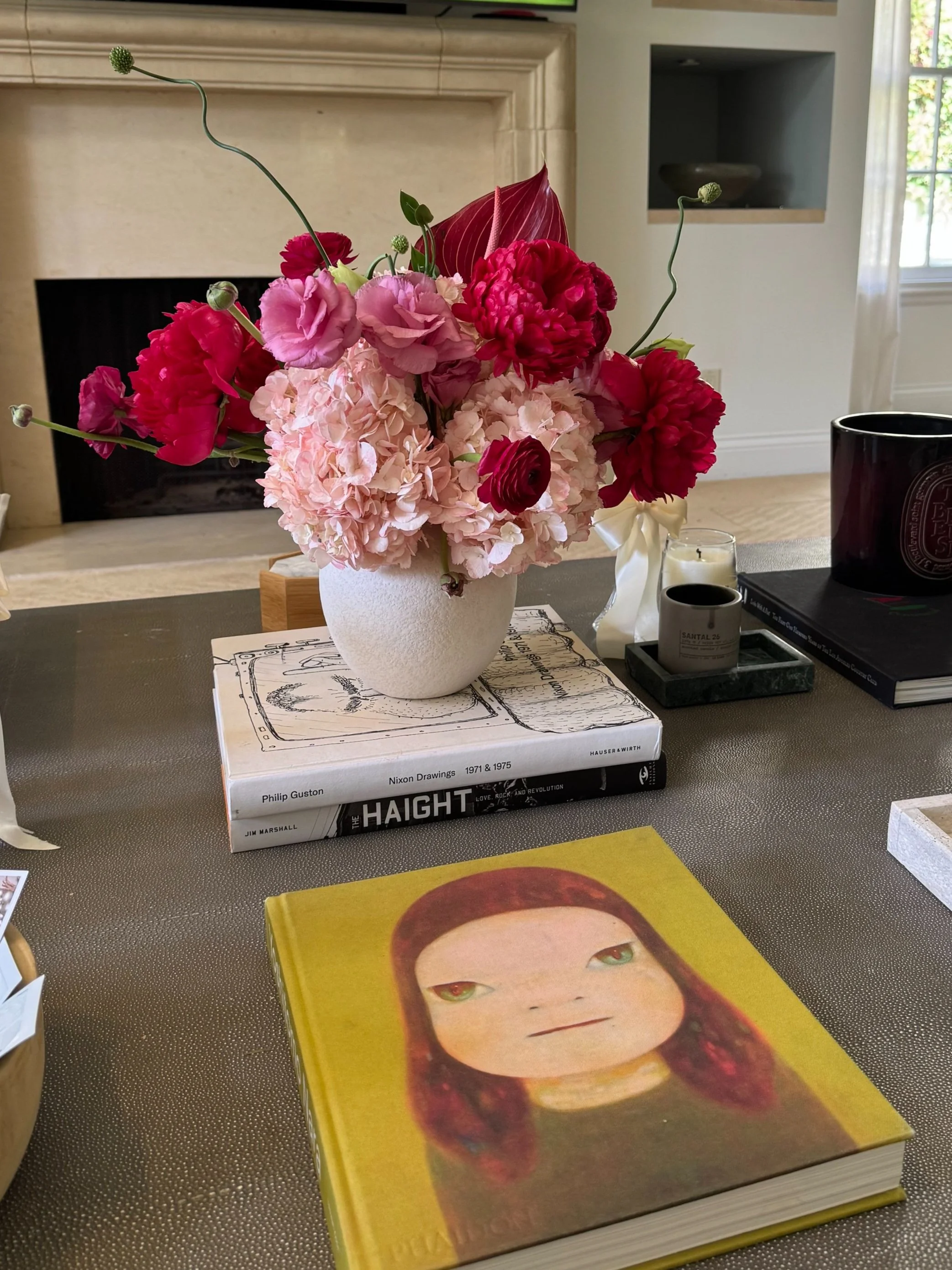 Vase of pink and red flowers on a table with books and a candle, in a room with fireplace and window.