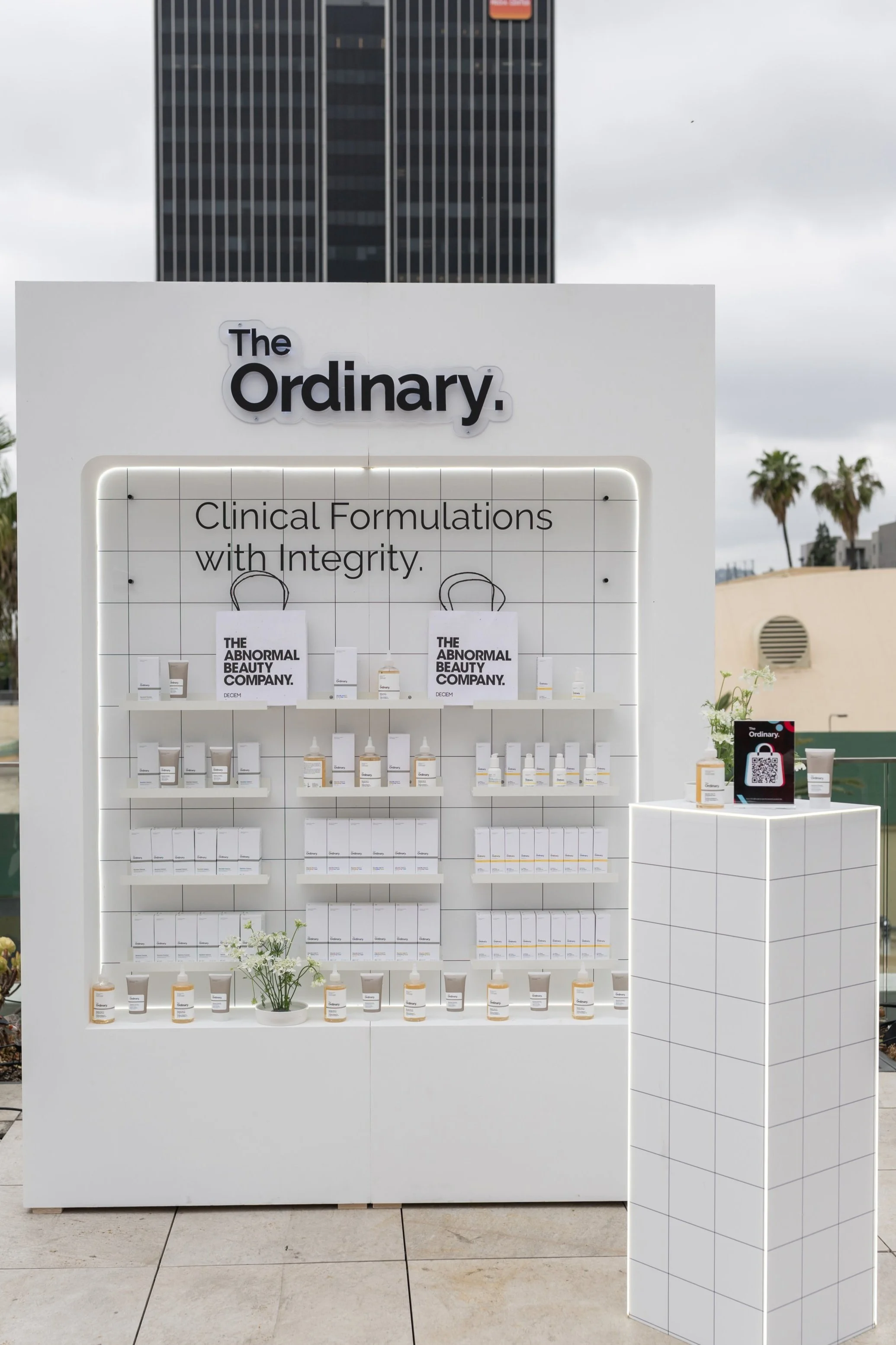 Display stand for The Ordinary skincare products with white minimalist design, featuring shelves with boxes and bottles, and signage stating 'Clinical Formulations with Integrity' and 'The Abnormal Beauty Company' at an outdoor event.