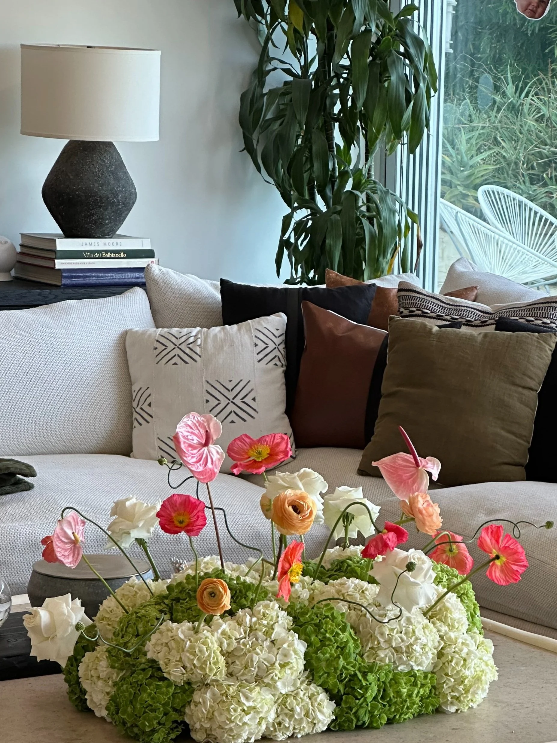 Living room with a cream-colored sofa, multiple decorative pillows, a large potted plant near a window, and a floral arrangement of white hydrangeas and pink, white, and peach flowers on a table.