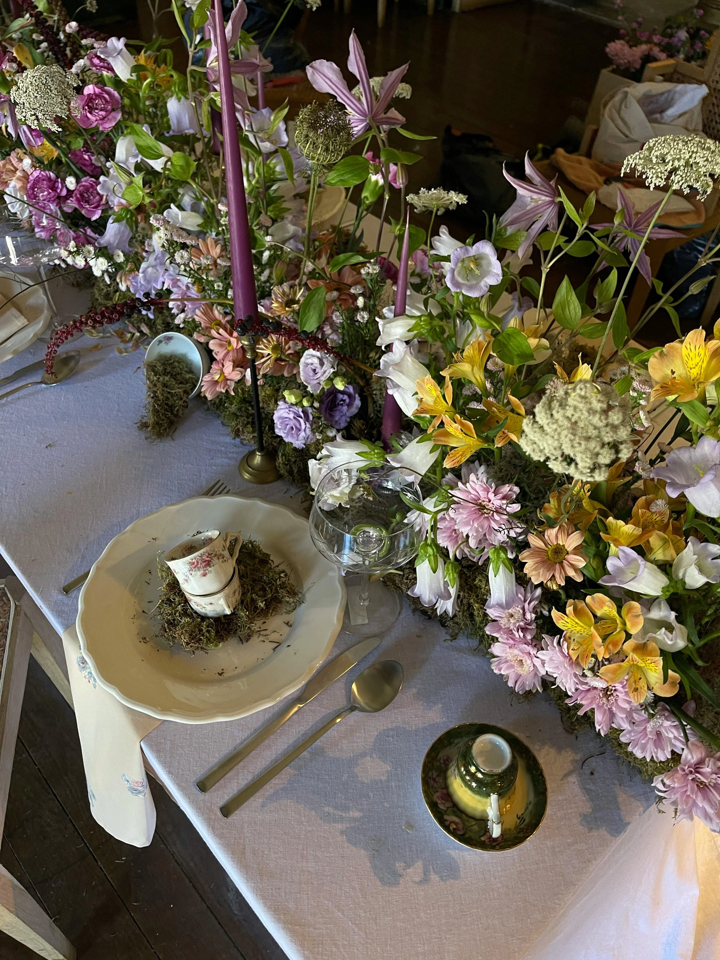 A table decorated with a large floral arrangement of pink, purple, yellow, and white flowers with candles, a tea cup with a floral pattern on a bed of moss, a glass bowl, and a small decorative item, set for a formal gathering.