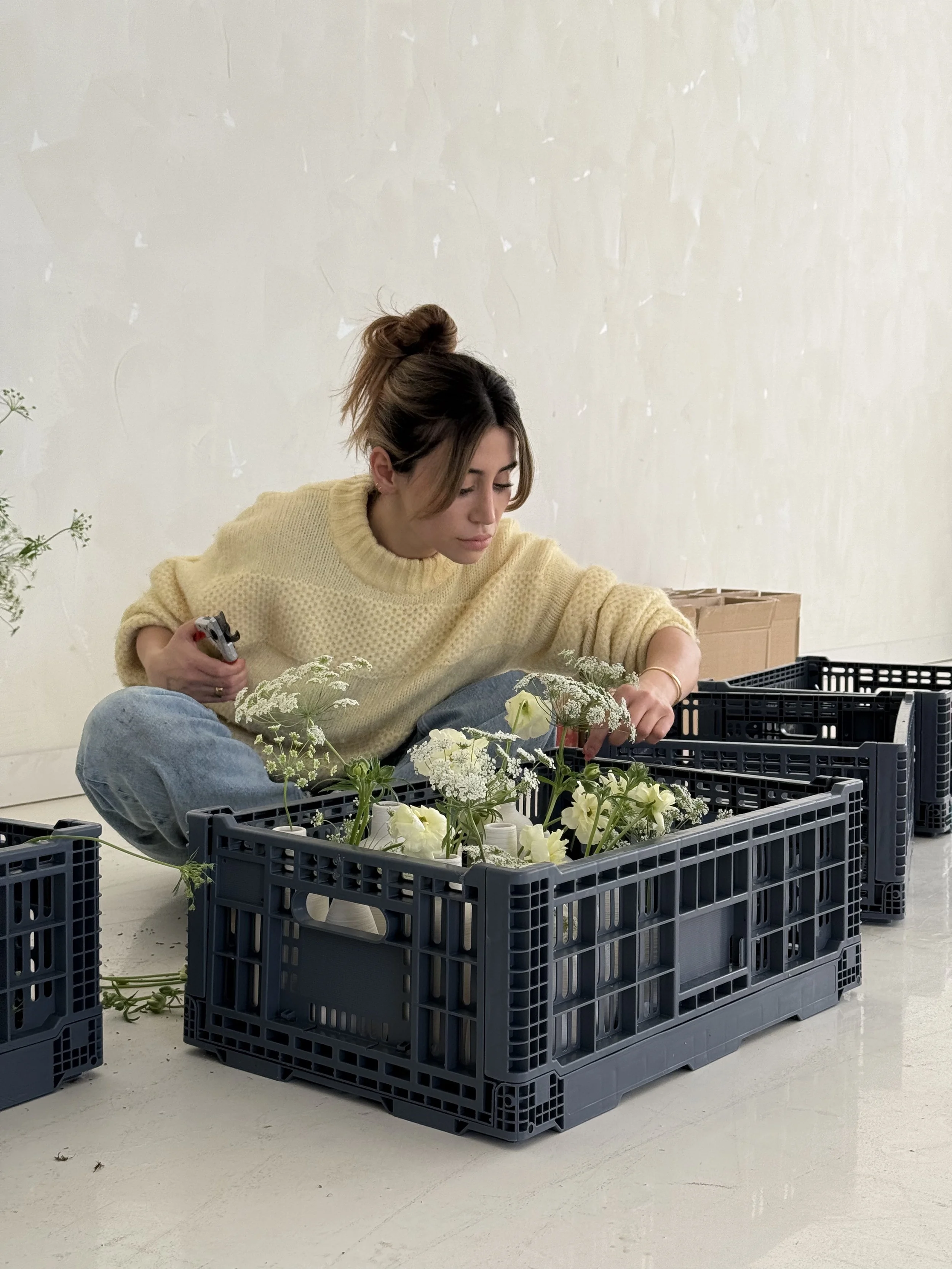 Woman arranging white flowers in black plastic crates, sitting on a floor in front of a plain wall.