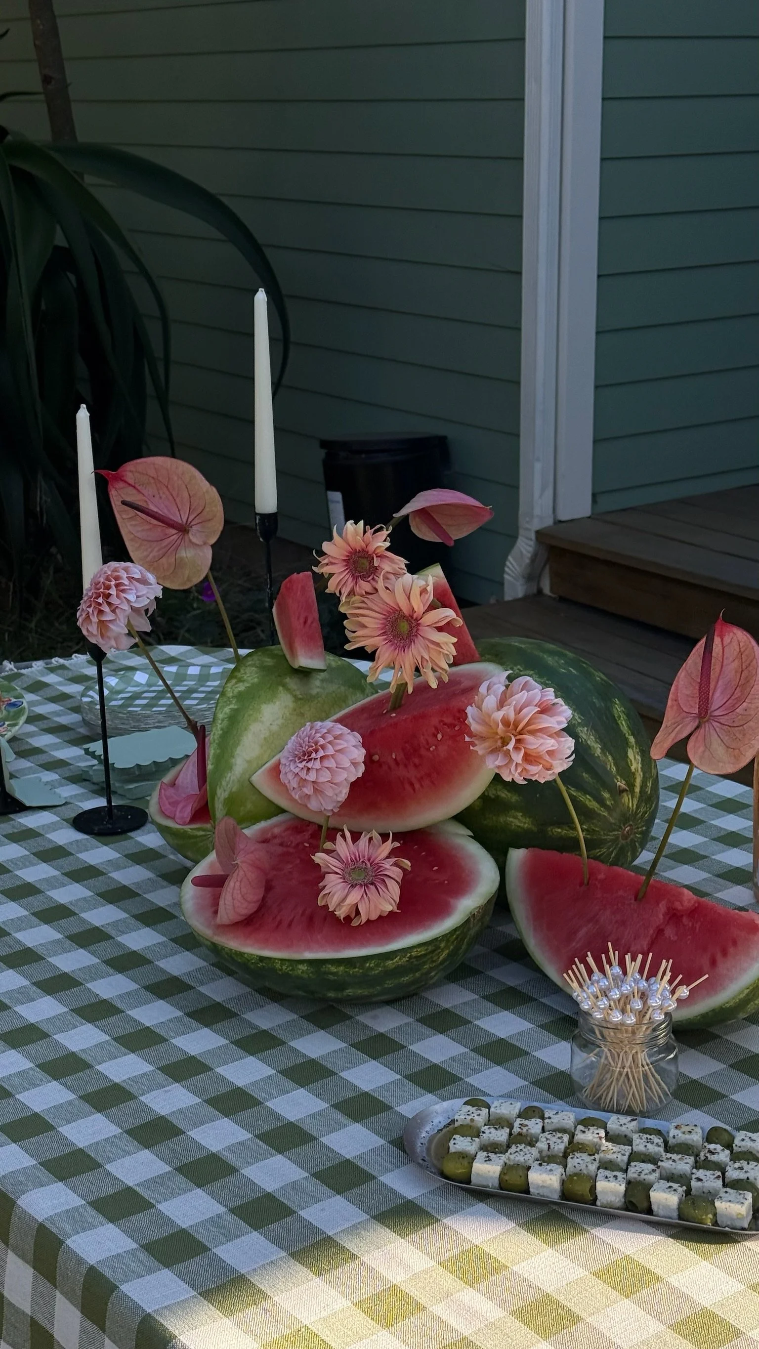 A table decorated with sliced watermelons, pink flowers, and pink anthuriums, with candles, toothpicks, and a tray of cheese and olives.