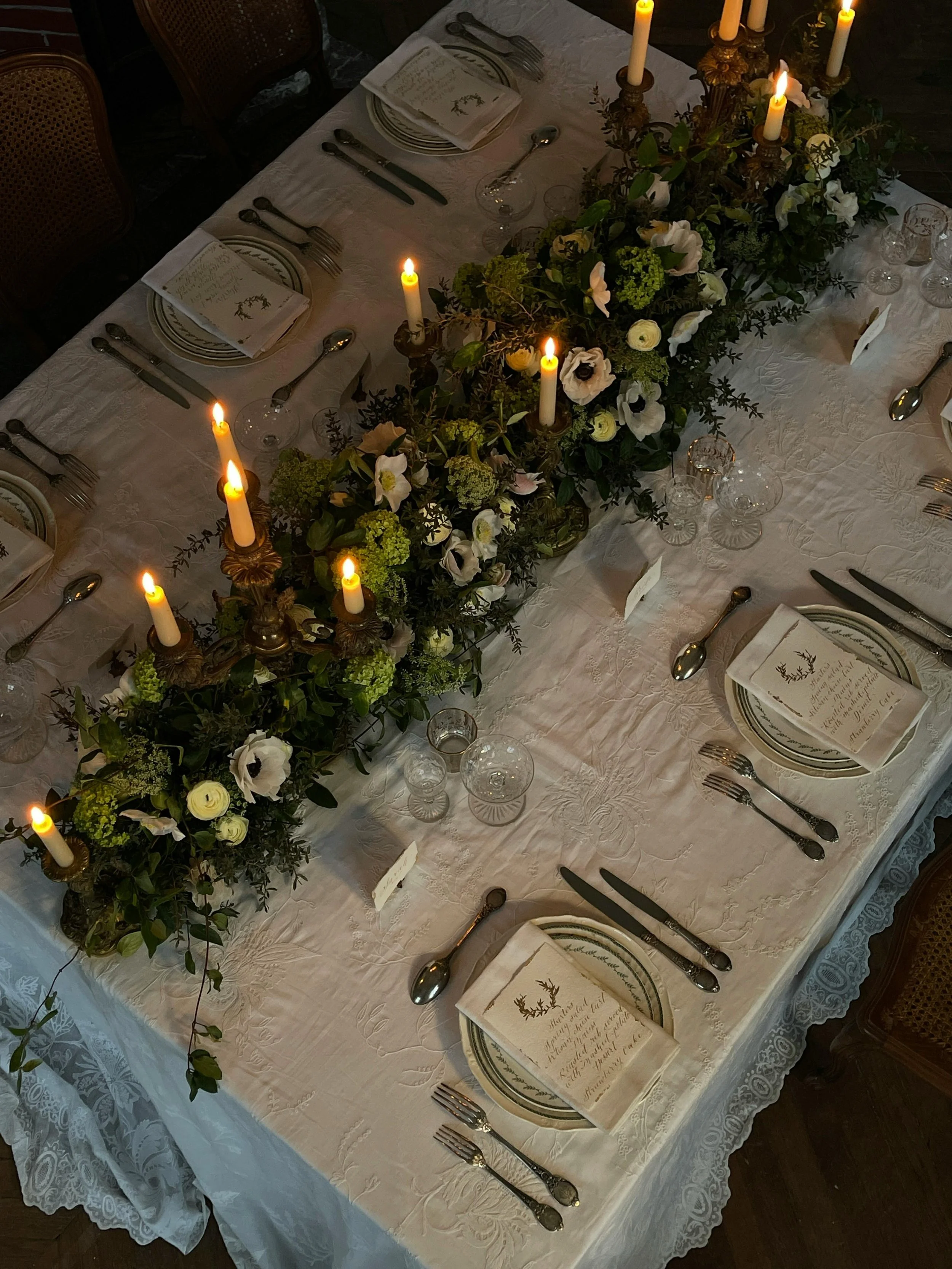 A long rectangular dining table set for a formal dinner with white tablecloth, decorative plates, silverware, glassware, and napkins, adorned with a lush green and white floral centerpiece and gold candelabras with lit candles.