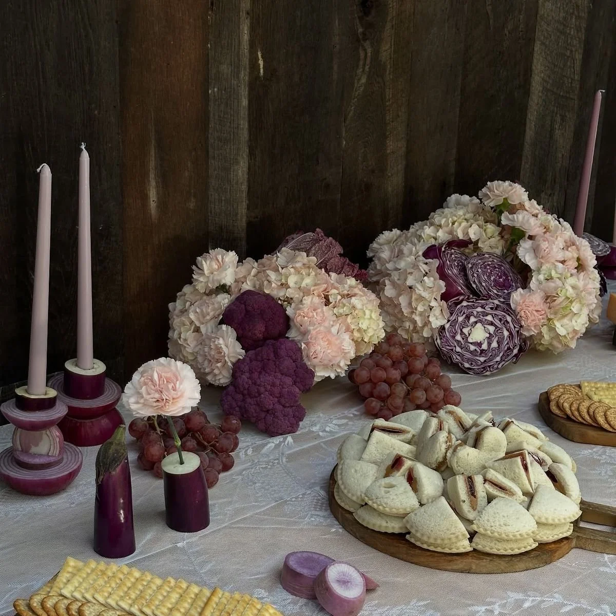 A table decorated with pink and purple flowers, grapes, cookies, and sandwiches on a white tablecloth, with two pink candles on the left side and a dark wooden background.