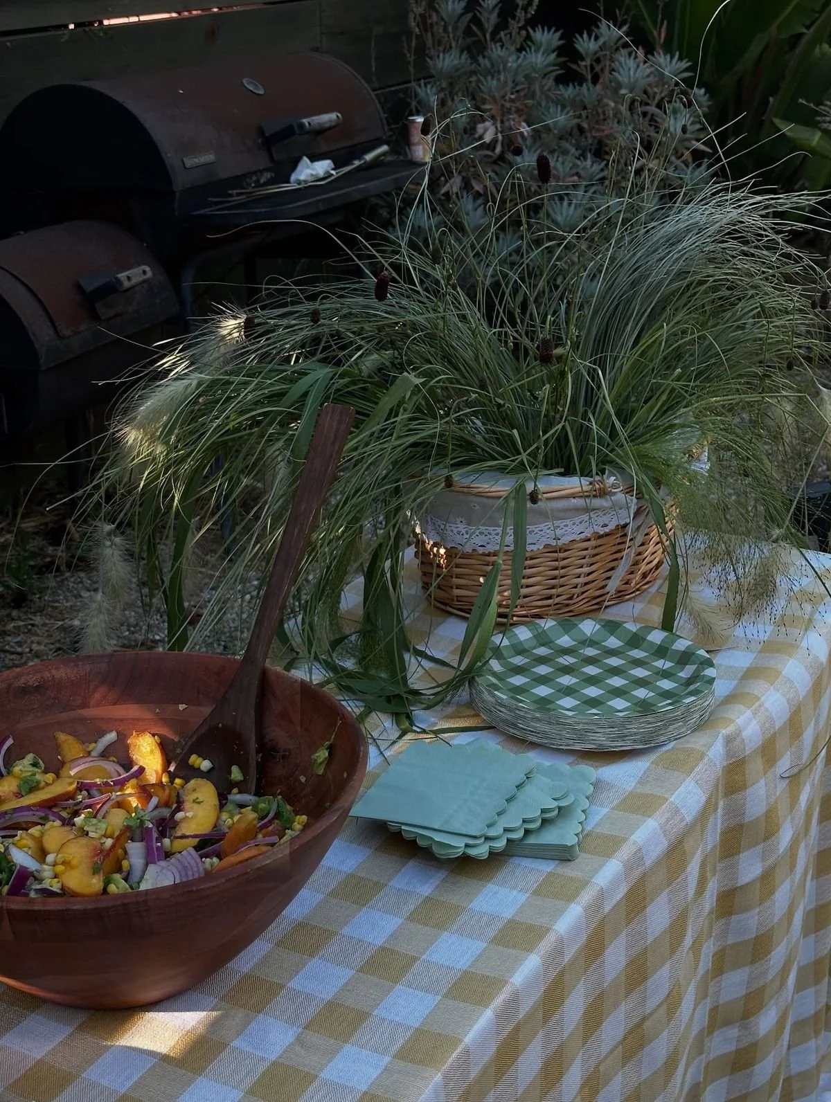 Table with a yellow and white checkered tablecloth, featuring a bowl of salad, disposable plates, napkins, a wicker basket with a cloth, and a large plant with tall green grasses.