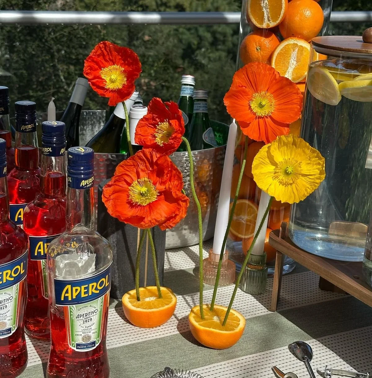 Table decorated with bright orange and yellow poppy flowers in orange halves, bottles of Aperol, glasses, and containers of sliced oranges and lemons at an outdoor gathering.