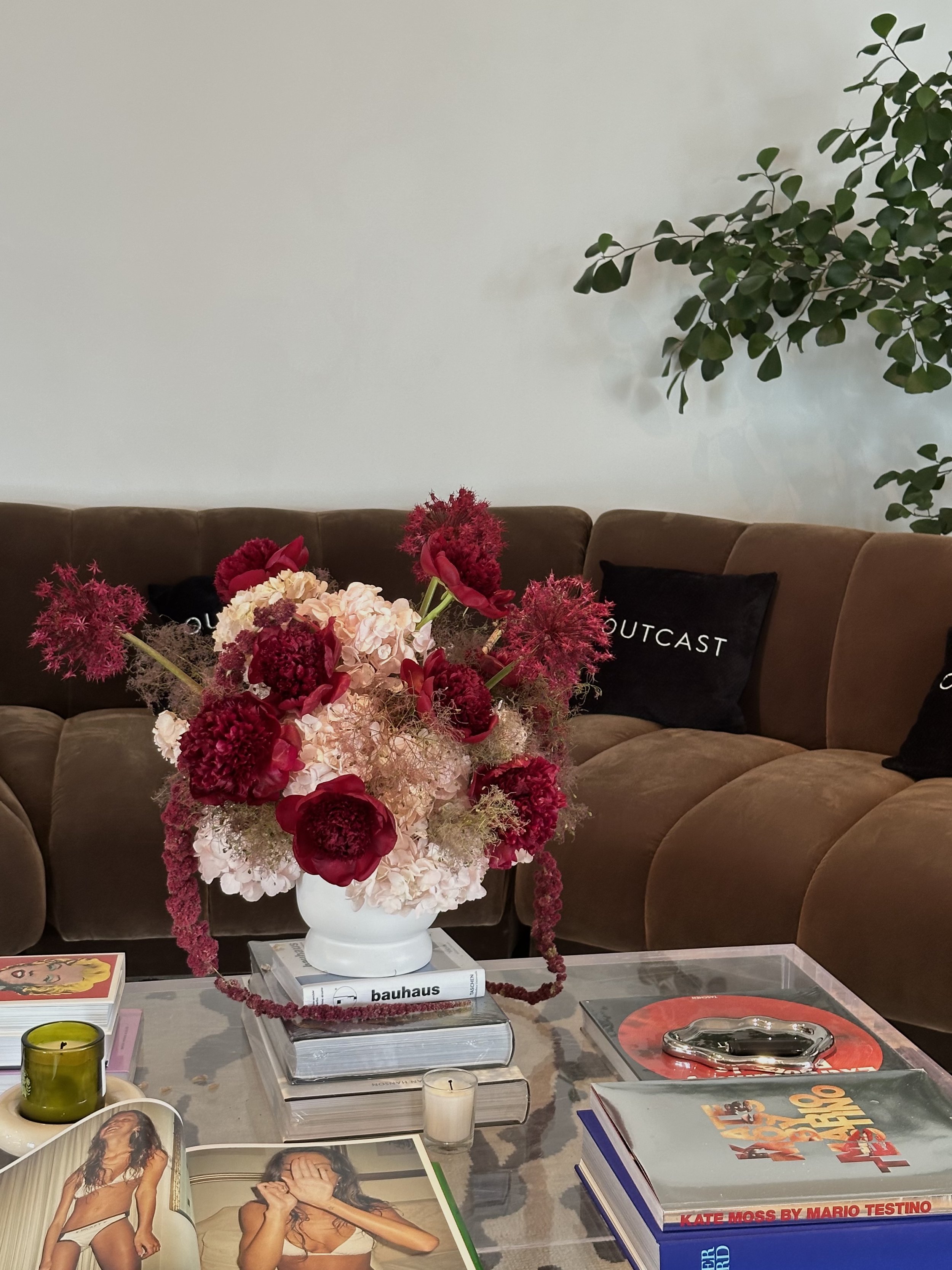 Living room with a tan sofa, a large floral arrangement of red, pink, and cream flowers in a white vase, and books on a glass table. Behind the sofa are black pillows with white text, and a green plant in the corner.