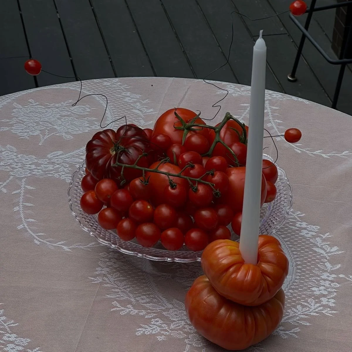 A crystal bowl filled with various tomatoes on a table, with a candle in a tomato-shaped holder. The tablecloth is light-colored with a textured pattern.