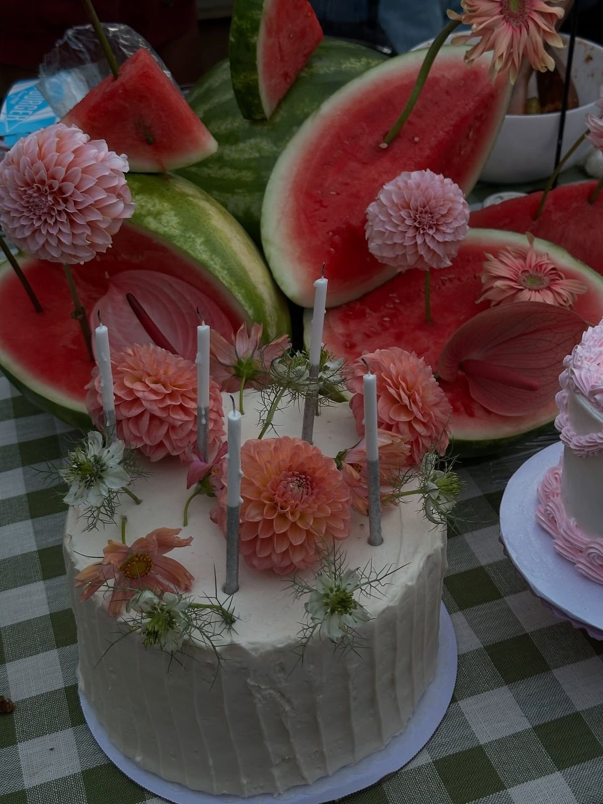 A celebration cake decorated with pink flowers and candles, surrounded by watermelon slices and additional pink flowers on a checkered tablecloth.