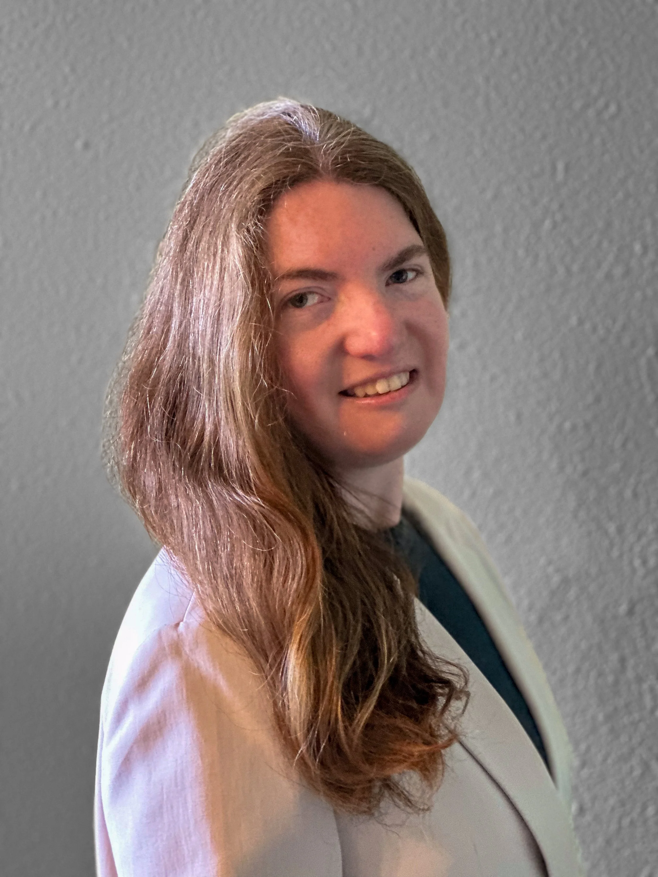 A woman with long brown hair, smiling, sitting in a modern office chair with a soft gray textured wall background.