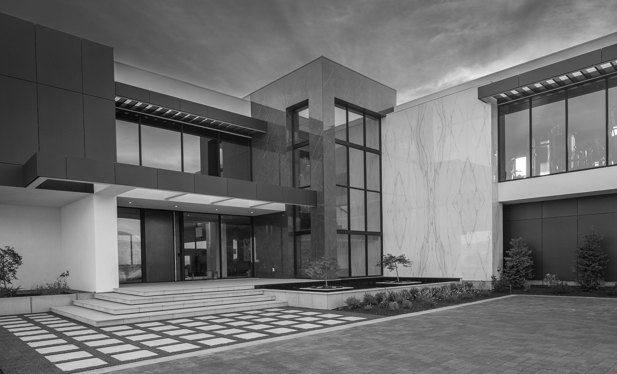 Modern architectural building with large glass windows, stairs leading to the entrance, and landscaped surroundings, in black and white.