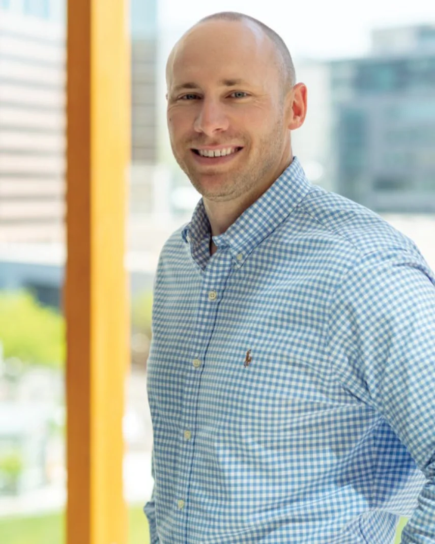 A man with a shaved head and blue eyes smiling, wearing a blue and white checkered button-up shirt, standing indoors near a large window with a cityscape background.