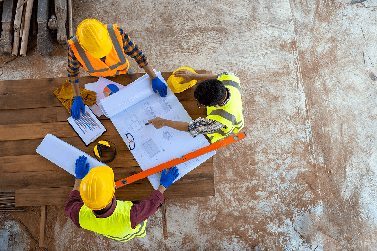 Three construction workers wearing yellow safety helmets and reflective vests examining blueprints on a wooden table, with construction tools and documents nearby, on a dusty construction site.