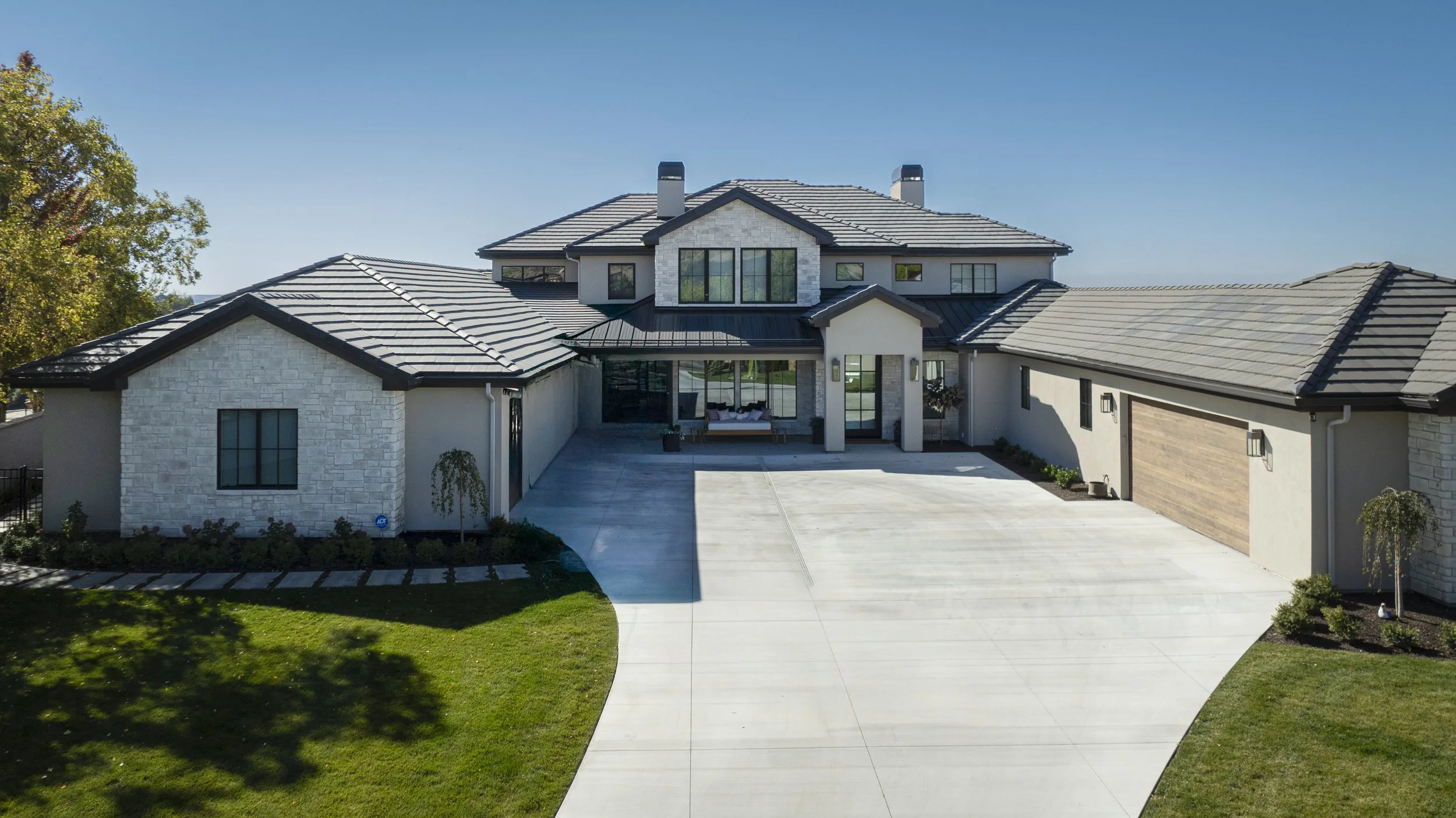 Front view of a modern two-story house with a light-colored exterior, dark roof, large windows, a spacious driveway, and neatly landscaped yard with green grass and small trees.