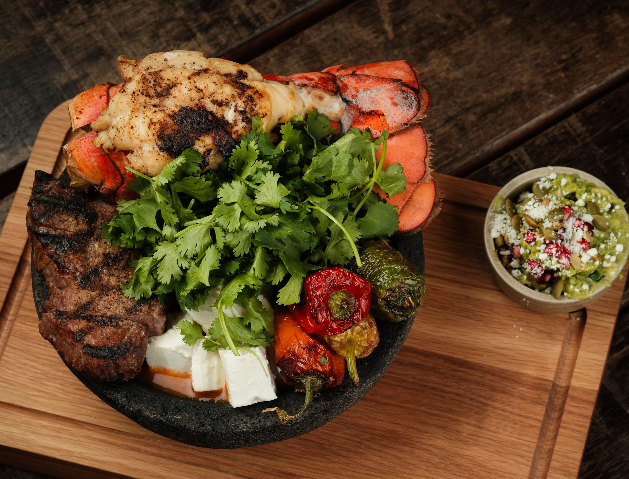 Lobster, cilantro, grilled peppers, grilled meat, cheese, and a side of guacamole with seeds and pico de gallo on a black stone plate on a wooden board.