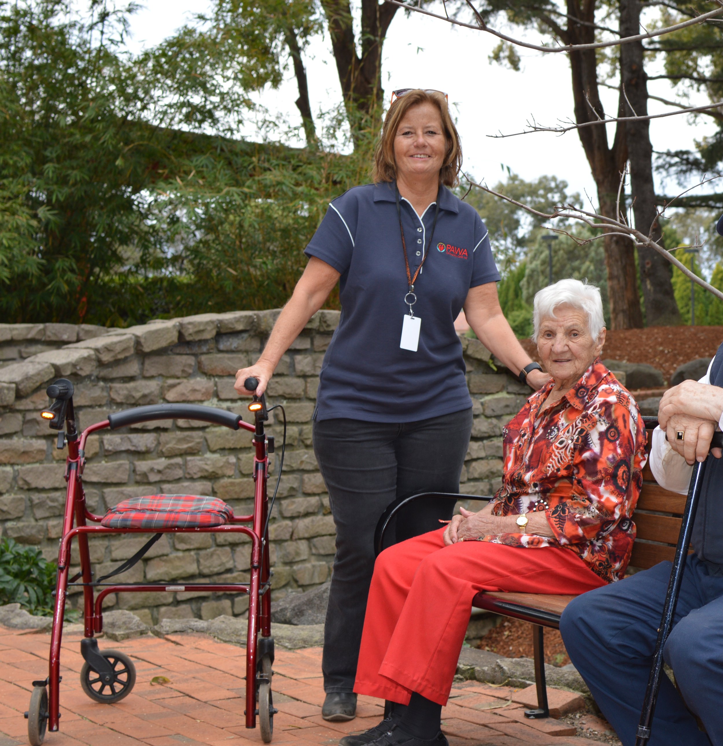 A woman with light skin, wearing a dark blue PAWA polo shirt, glasses on head, smiling while standing outdoors next to an elderly woman seated on a bench, surrounded by greenery and trees.