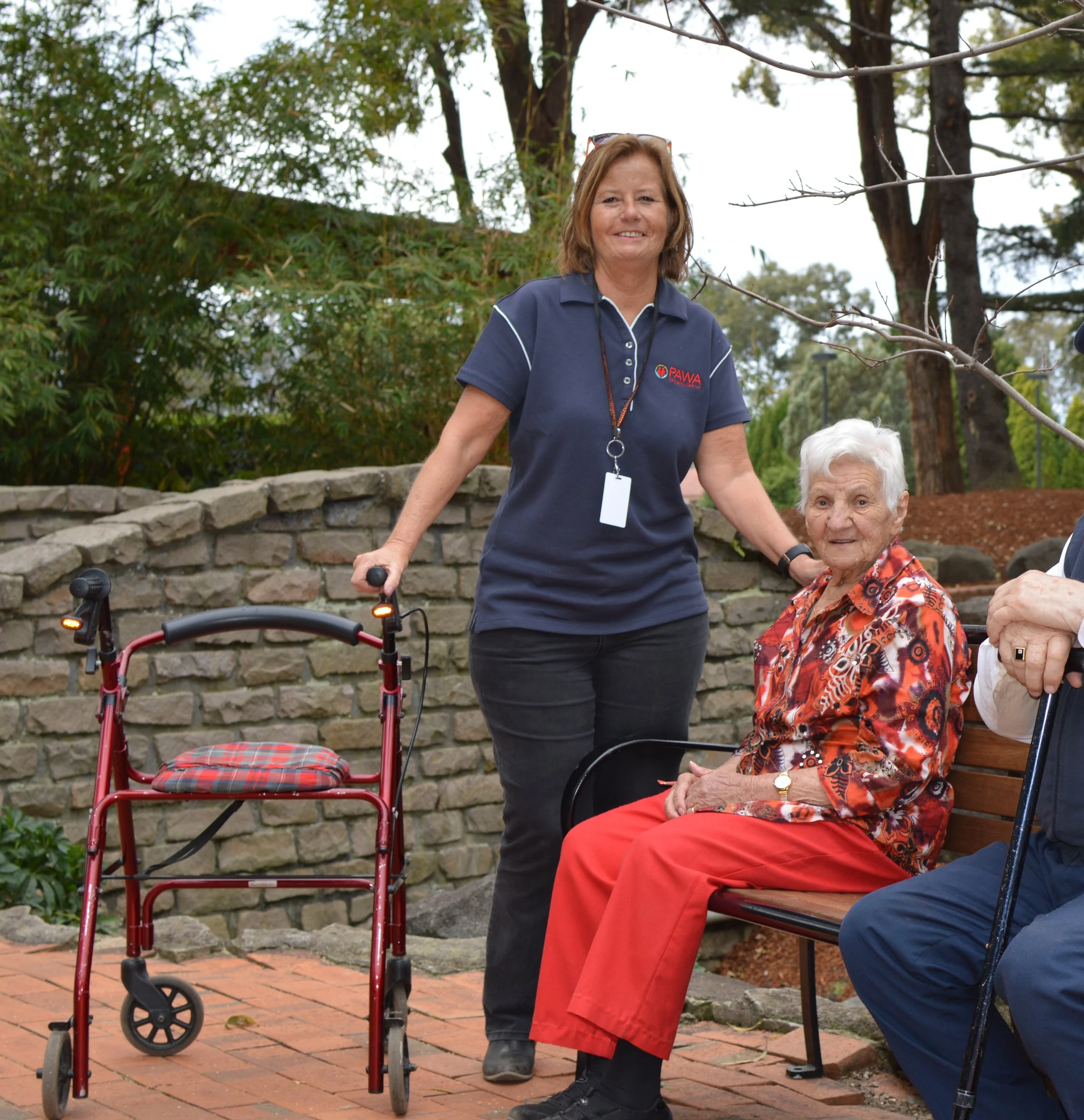 A woman with light skin, wearing a dark blue PAWA polo shirt, glasses on head, smiling while standing outdoors next to an elderly woman seated on a bench, surrounded by greenery and trees.
