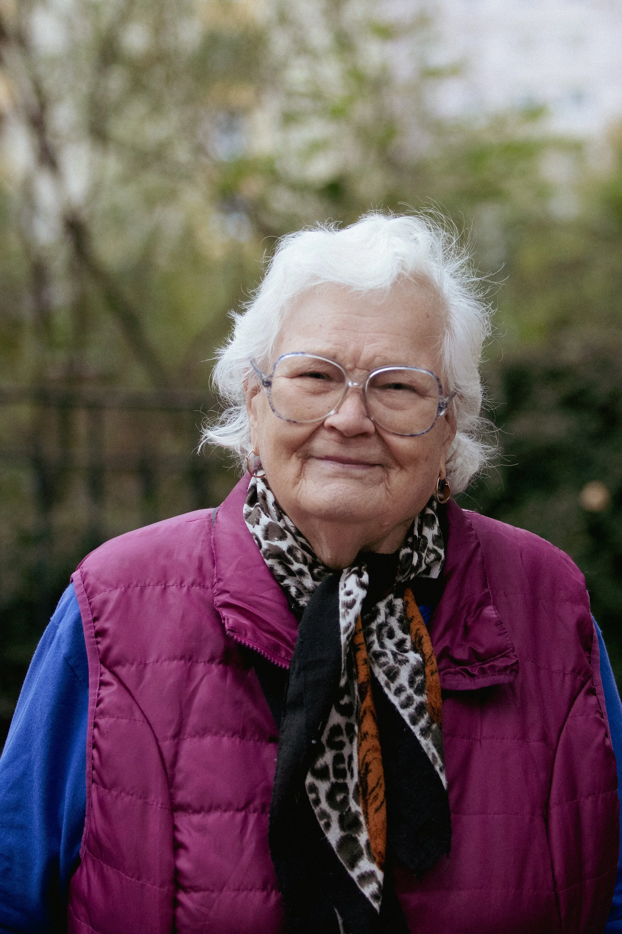 An elderly woman with white hair, glasses, wearing a purple vest over a blue shirt, and a leopard print scarf, standing outdoors with blurred trees in the background.