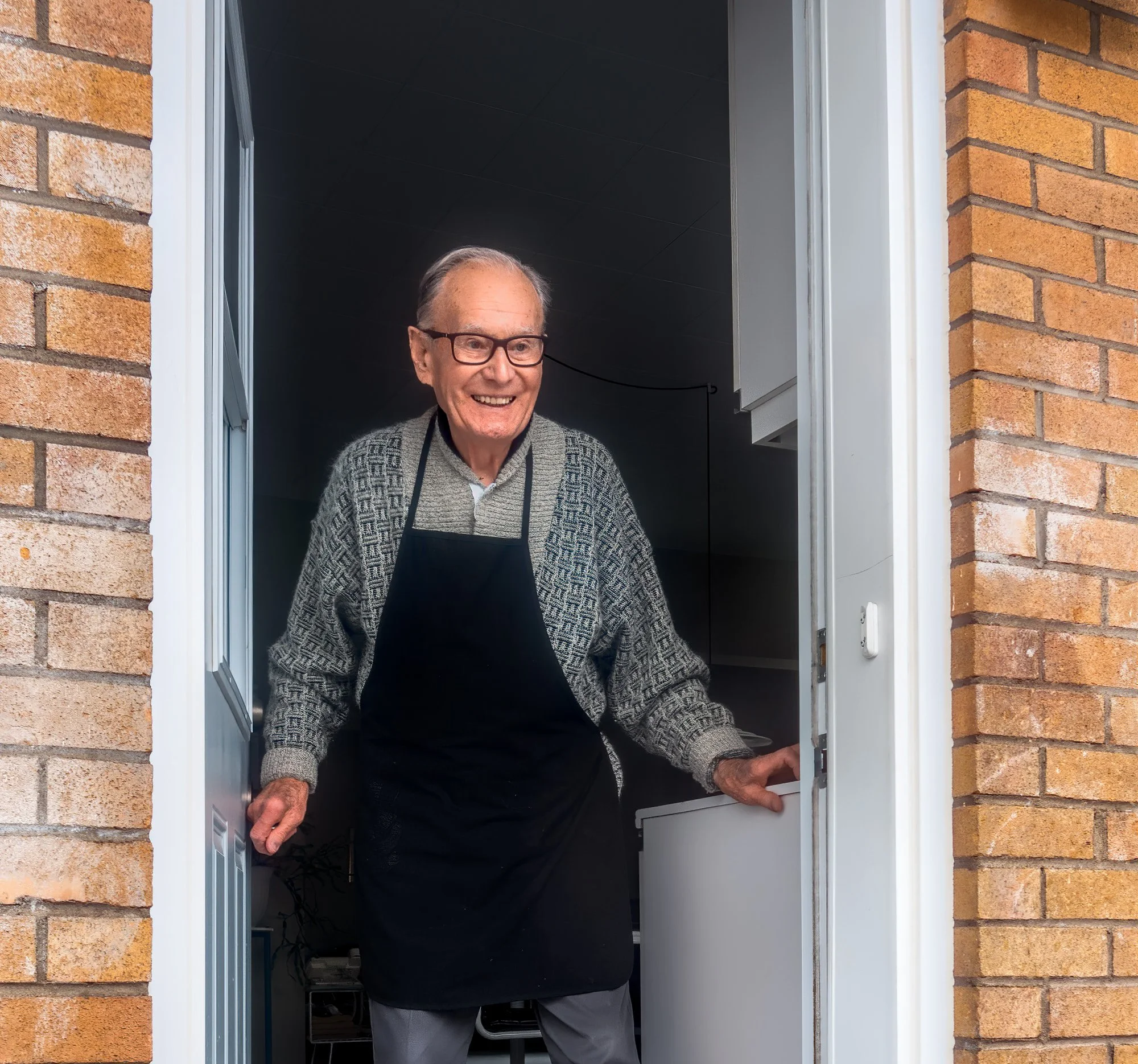 An elderly man with glasses smiling and waving at the doorway of a house.