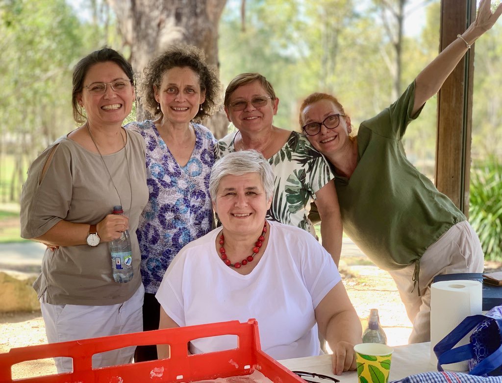 Five women smiling outdoors, with one seated at a table and the other four standing behind office