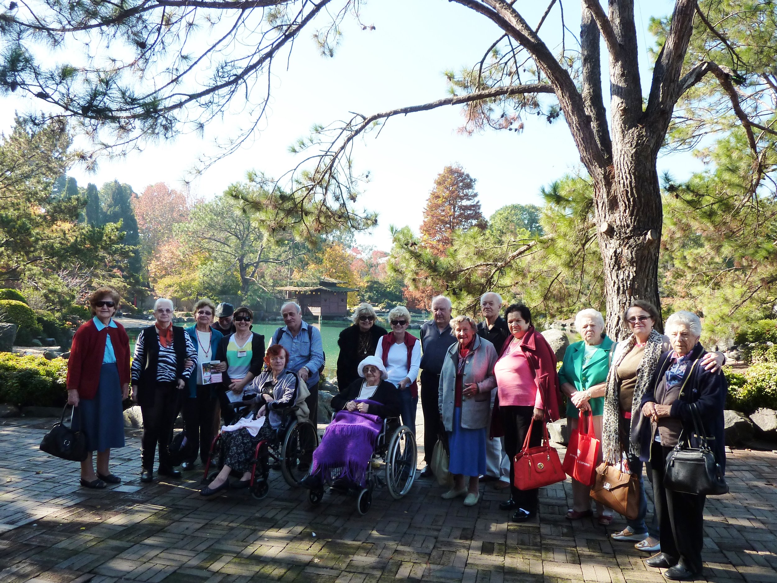 A group of elderly people, some in wheelchairs, standing outdoors in a garden with large trees and colorful foliage in the background.
