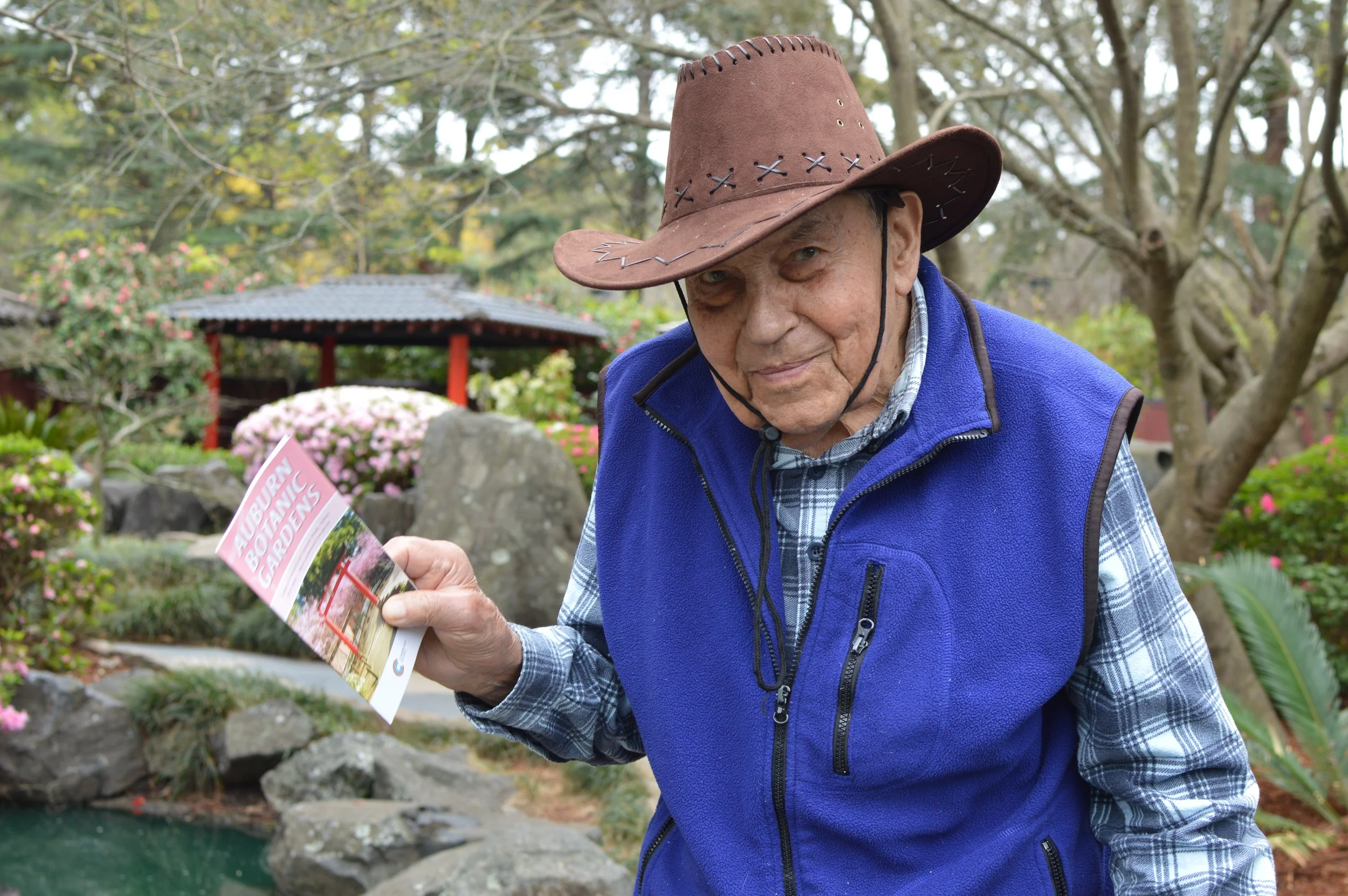 An elderly man wearing a brown cowboy hat and blue vest reading a brochure titled 'Autumn Botanical Gardens' outdoors in a garden with trees, blooming flowers, and a small pond in the background.