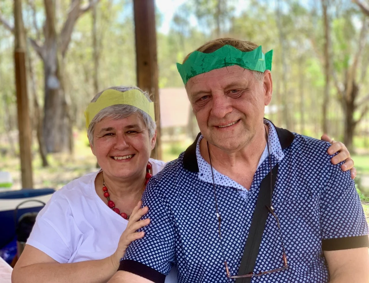 A smiling woman and a smiling man wearing paper crowns, outdoors in a park-like setting.