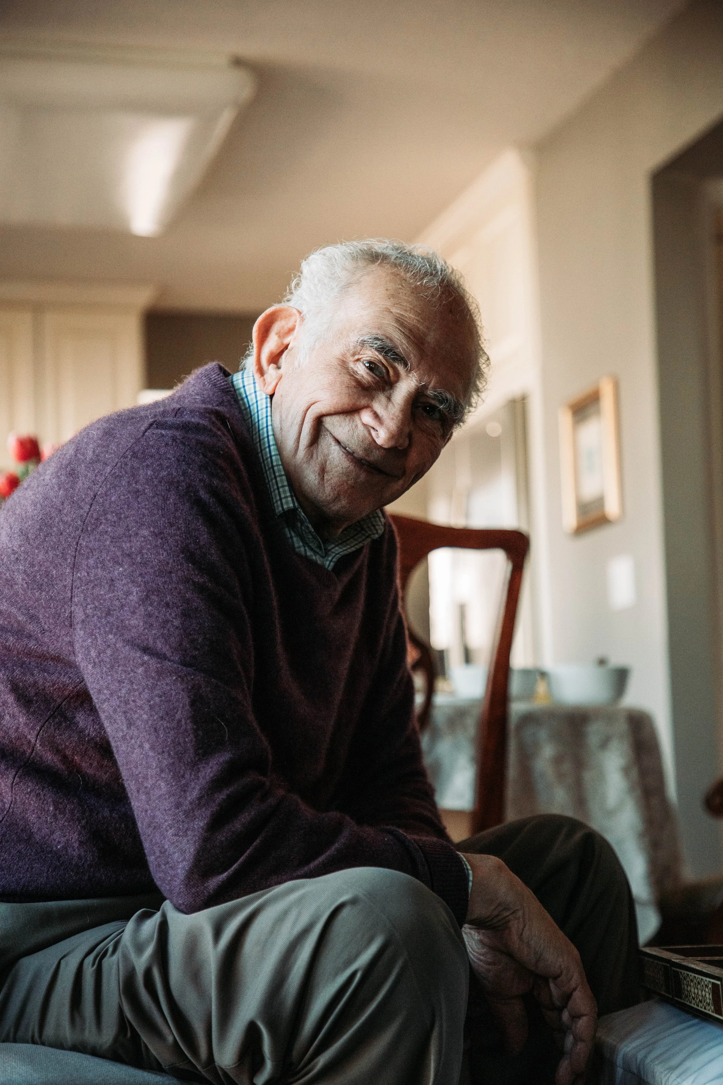 An elderly man with gray hair, wearing a purple sweater and checkered shirt, smiling and leaning forward indoors. The background shows a dining area with a table, a chair, framed artwork on the wall, and curtains.