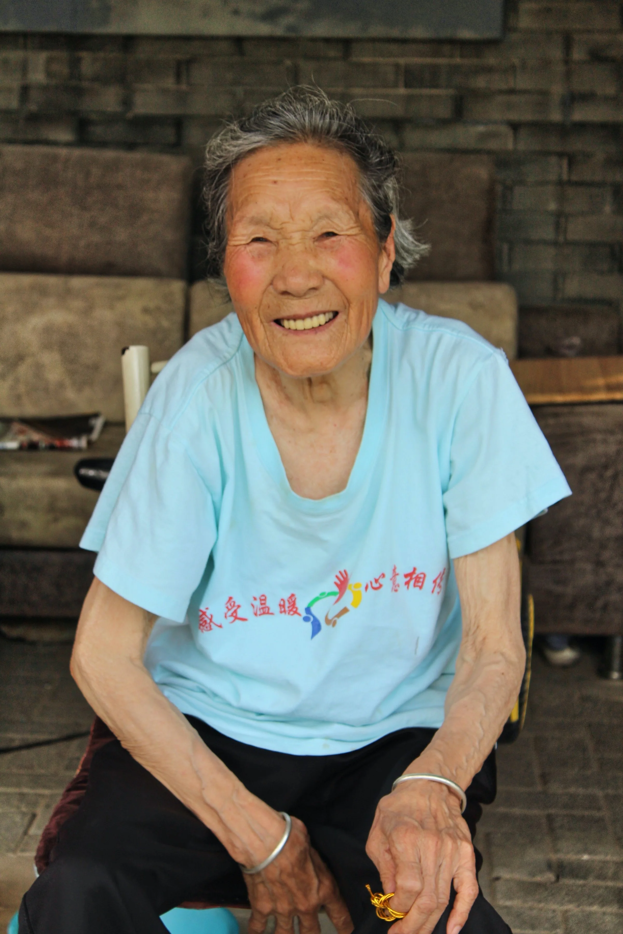 An elderly woman smiling while sitting on a chair against a rustic brick wall. She has gray hair, wearing a light blue T-shirt with colorful Chinese characters and symbols, black pants, and accessories including a silver bracelet and gold rings.