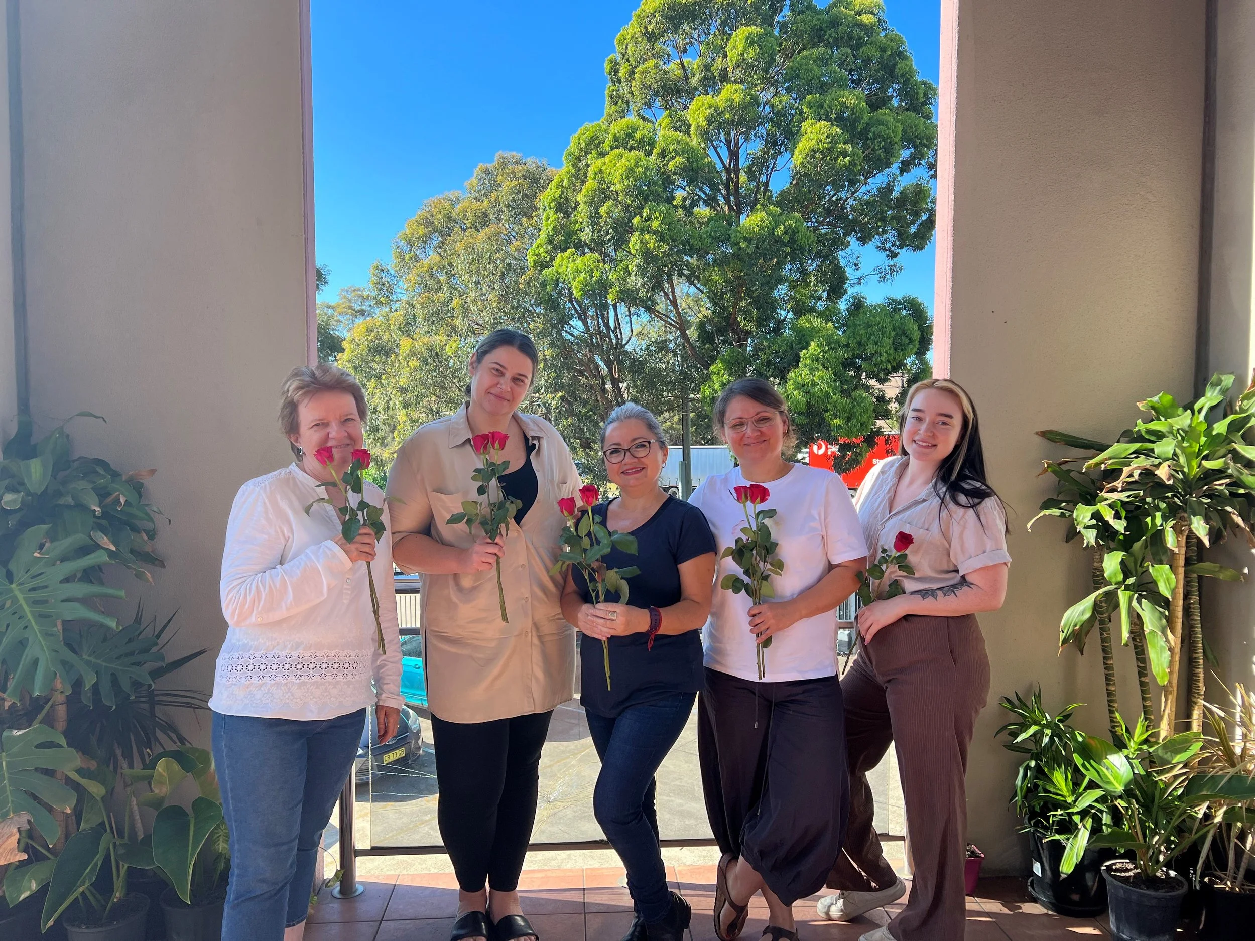 Five women standing together in a doorway, holding pink roses and smiling. Lush green trees and a blue sky are visible outside.