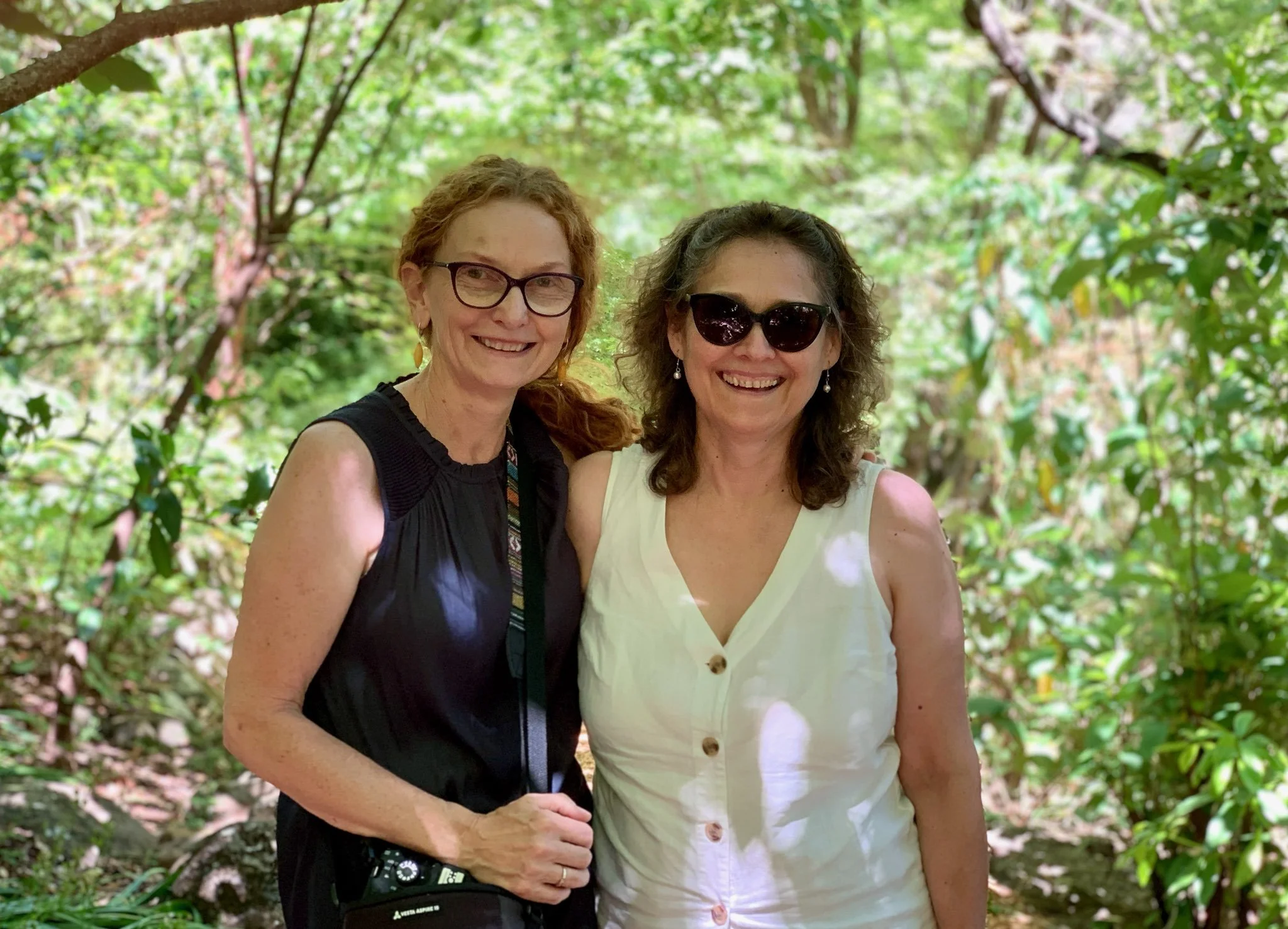 Two women smiling and standing close together outdoors in a lush green wooded area.