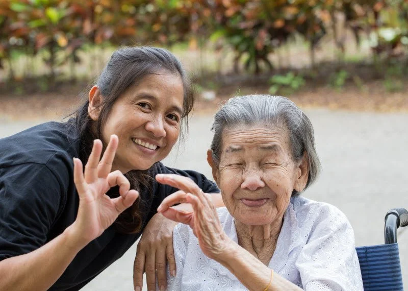 A young woman and an elderly woman smiling and making an OK sign together outdoors